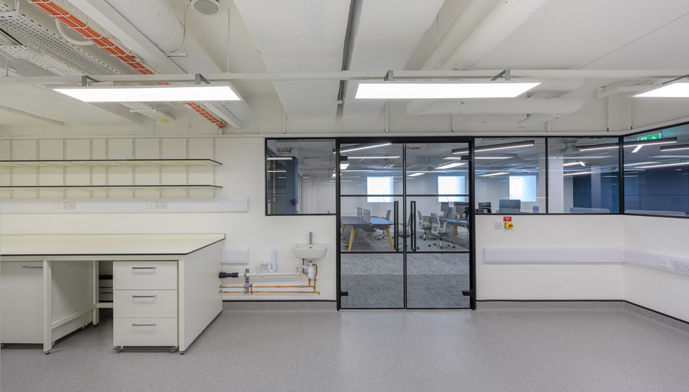Clean laboratory with white workstations, open shelving, and a glass-partitioned office area in the background.
