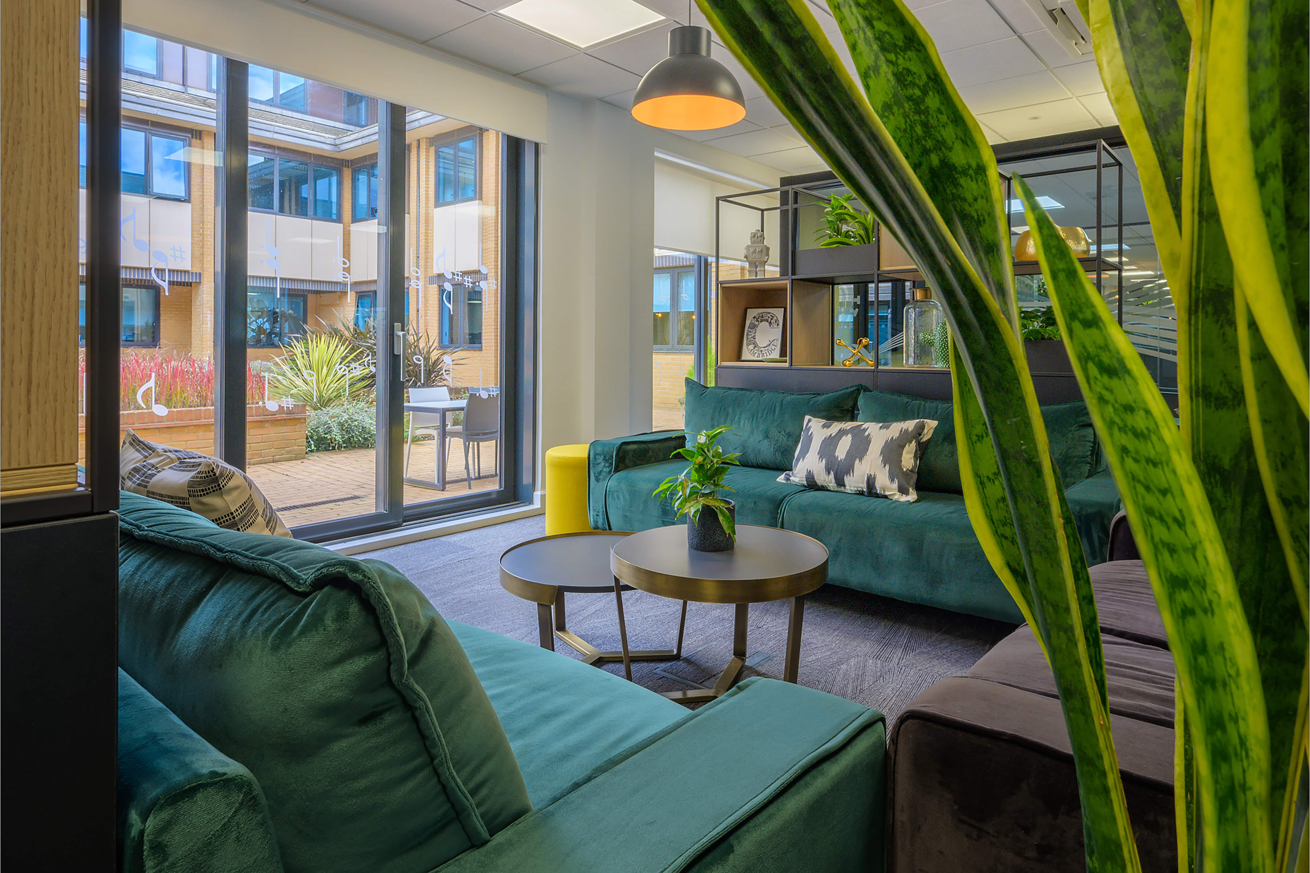Modern office lounge with green sofas, a black coffee table, and indoor plants. Large windows offer views of an outdoor courtyard. Calm ambiance
