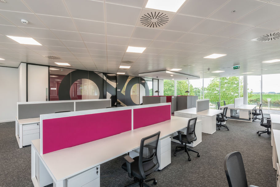 Modern office space with a row of white desks and pink dividers, black chairs, and large windows revealing a green landscape. Bright and open atmosphere