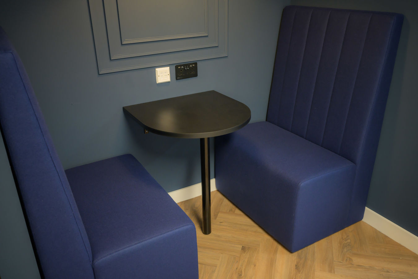 Blue upholstered booth seating with a small black table in the center against a blue paneled wall; a minimalist, cozy, and modern setting.