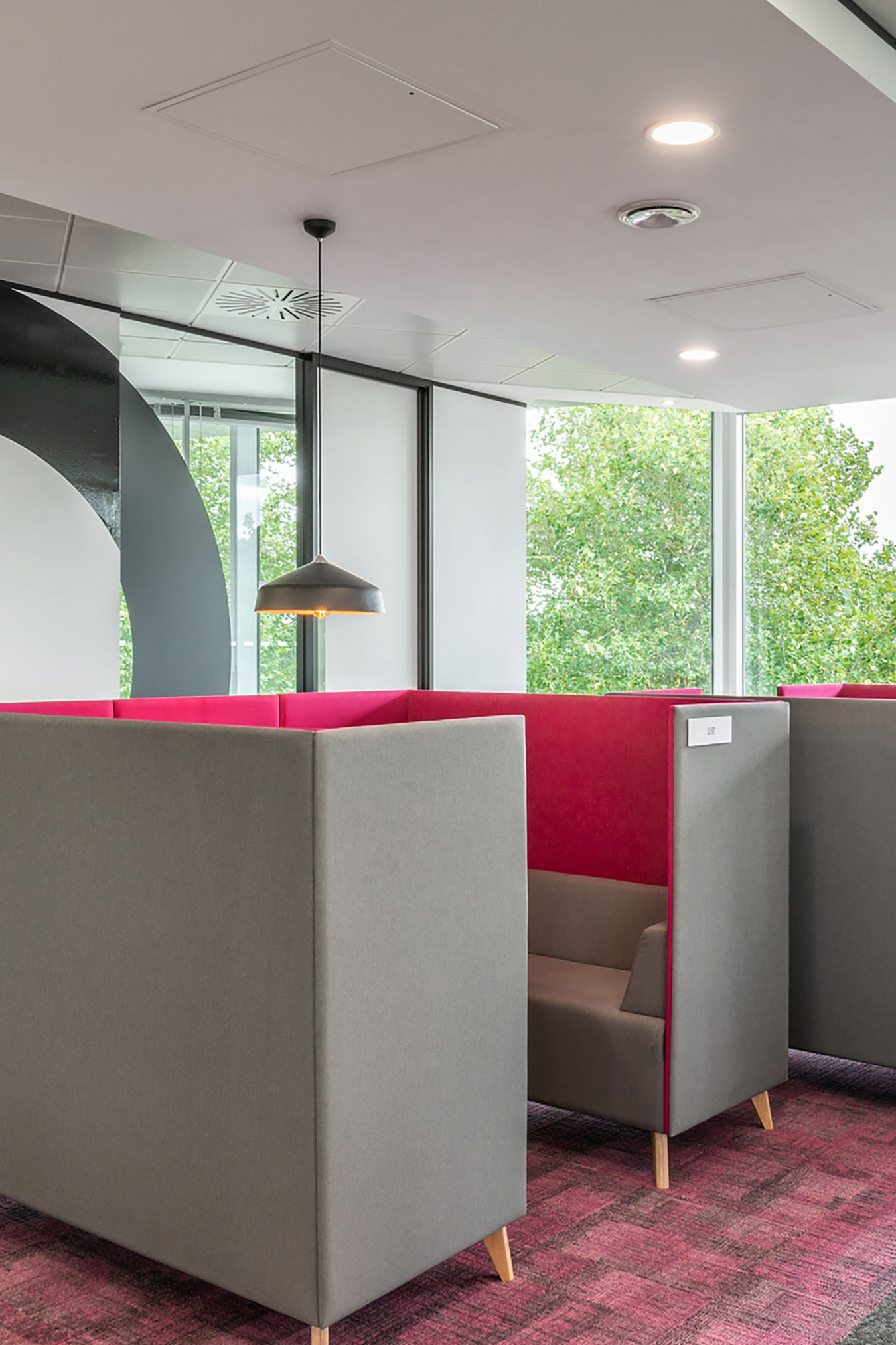 Modern office space with cozy gray booths featuring vibrant pink interiors, set on a patterned carpet. Large windows display green trees outside