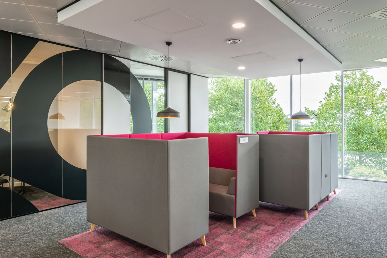 Modern office space with cozy gray booths featuring vibrant pink interiors, set on a patterned carpet. Large windows display green trees outside