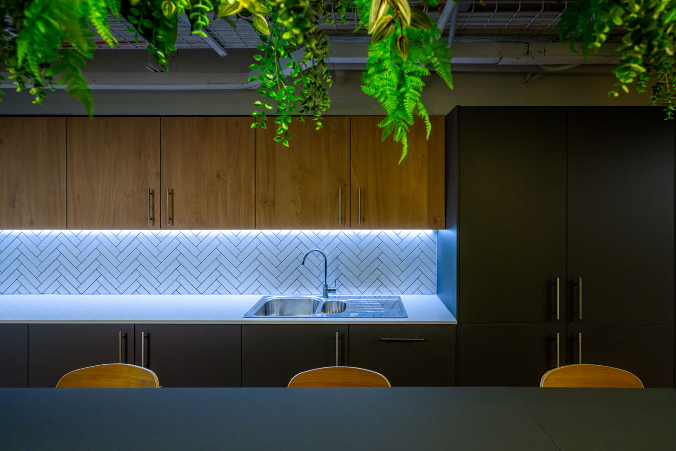 Kitchen sink with a sleek faucet is installed on a countertop beneath herringbone-tiled backsplash and wooden cabinets, surrounded by hanging green plants.
