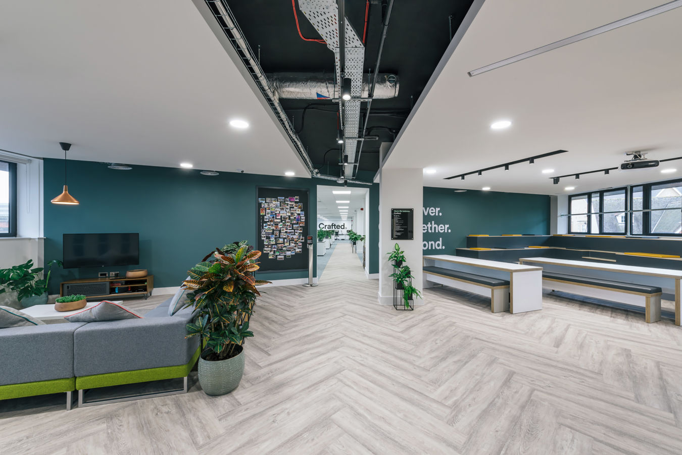 Modern office space with teal walls, exposed ductwork, and herringbone floors. Features include a seating area, plants, a TV, and tiered benches, conveying a casual, collaborative vibe.