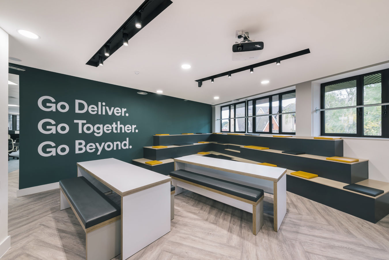 Modern office space with tiered seating in gray and yellow, large windows, and motivational phrases on a dark wall. Bright and open atmosphere