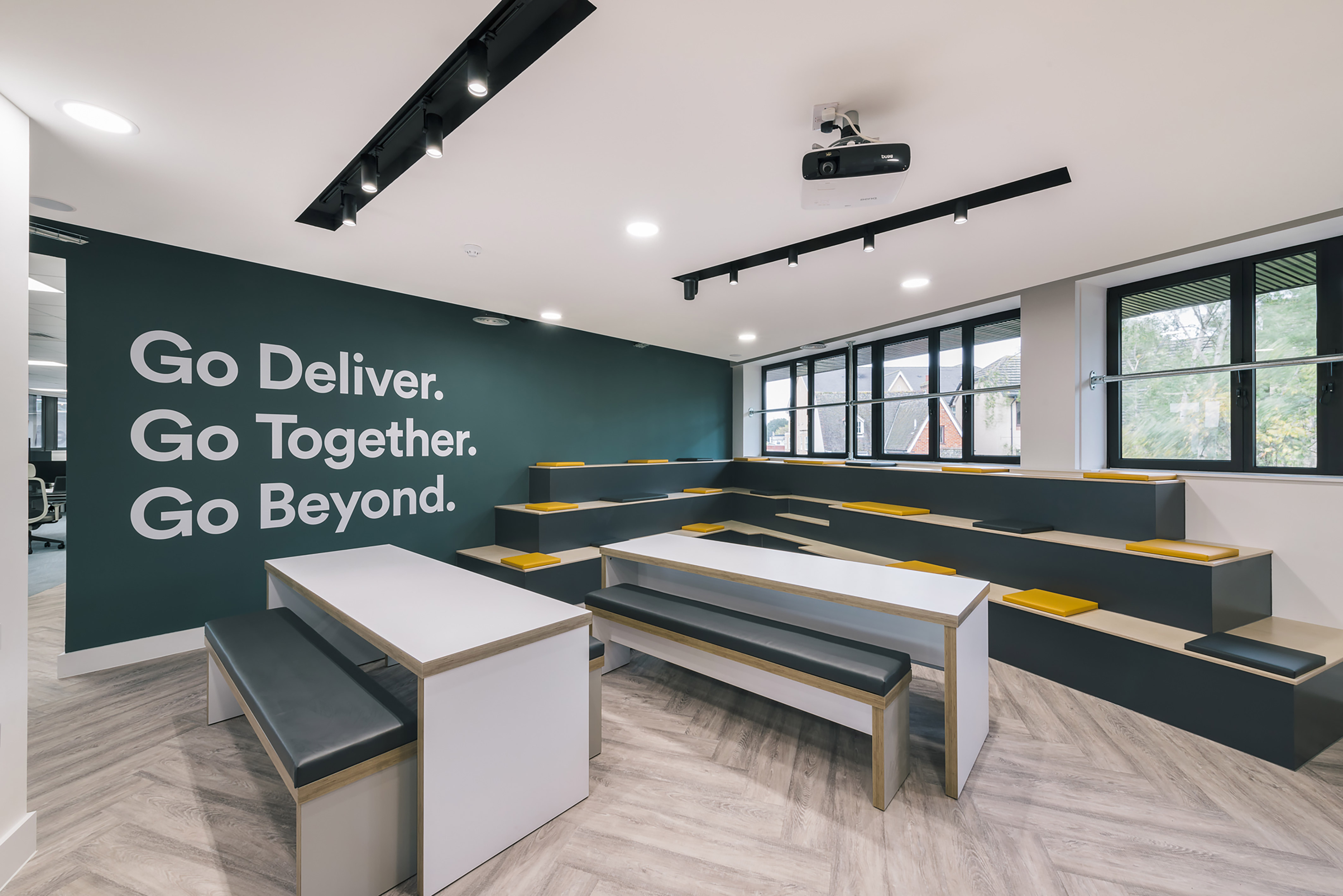 Modern office space with tiered seating in gray and yellow, large windows, and motivational phrases on a dark wall. Bright and open atmosphere