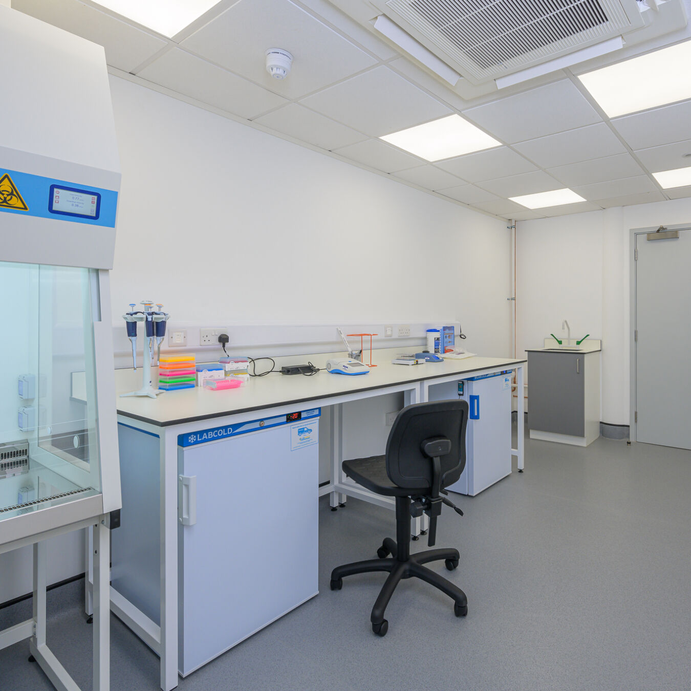 Clean laboratory room with a workbench, lab equipment, fume hood, and an adjustable stool under bright ceiling lights