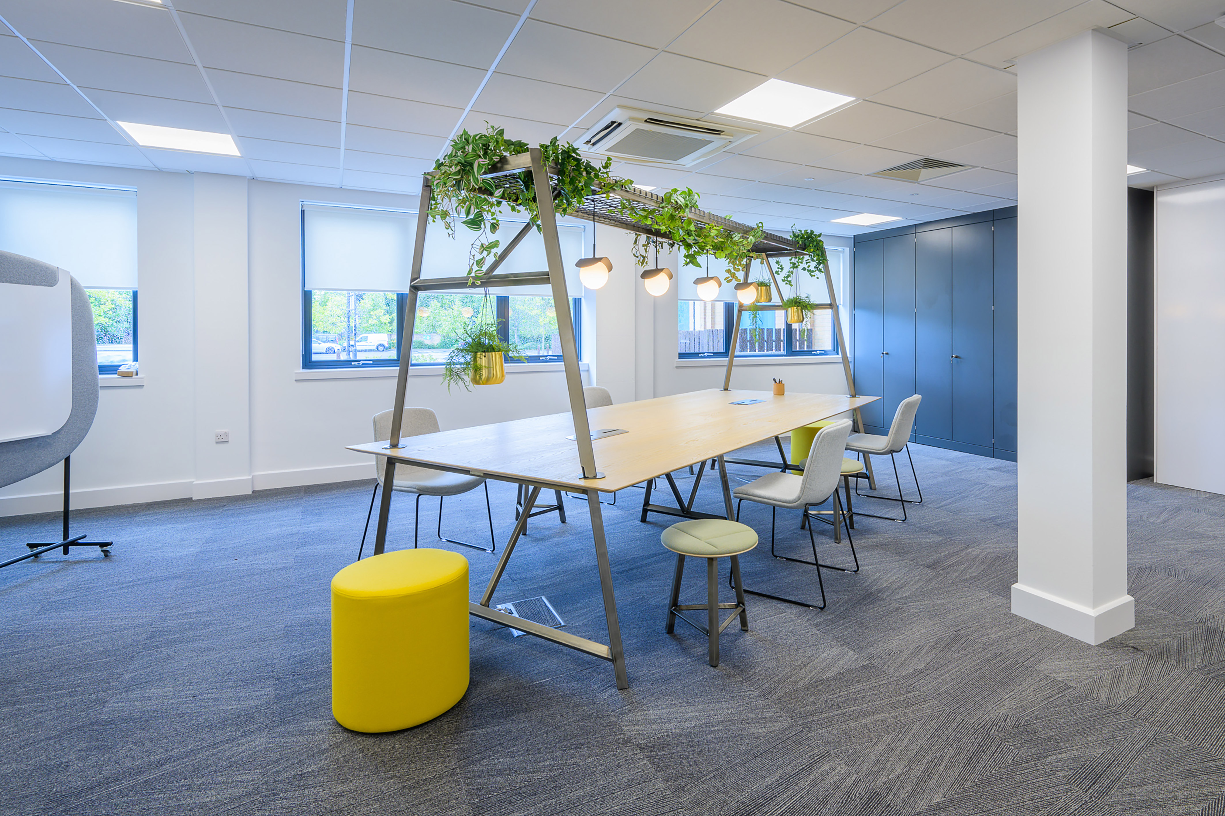 Large conference table decorated with hanging plants and lights, surrounded by chairs and a yellow stool in a bright office space with carpeted floor and windows.