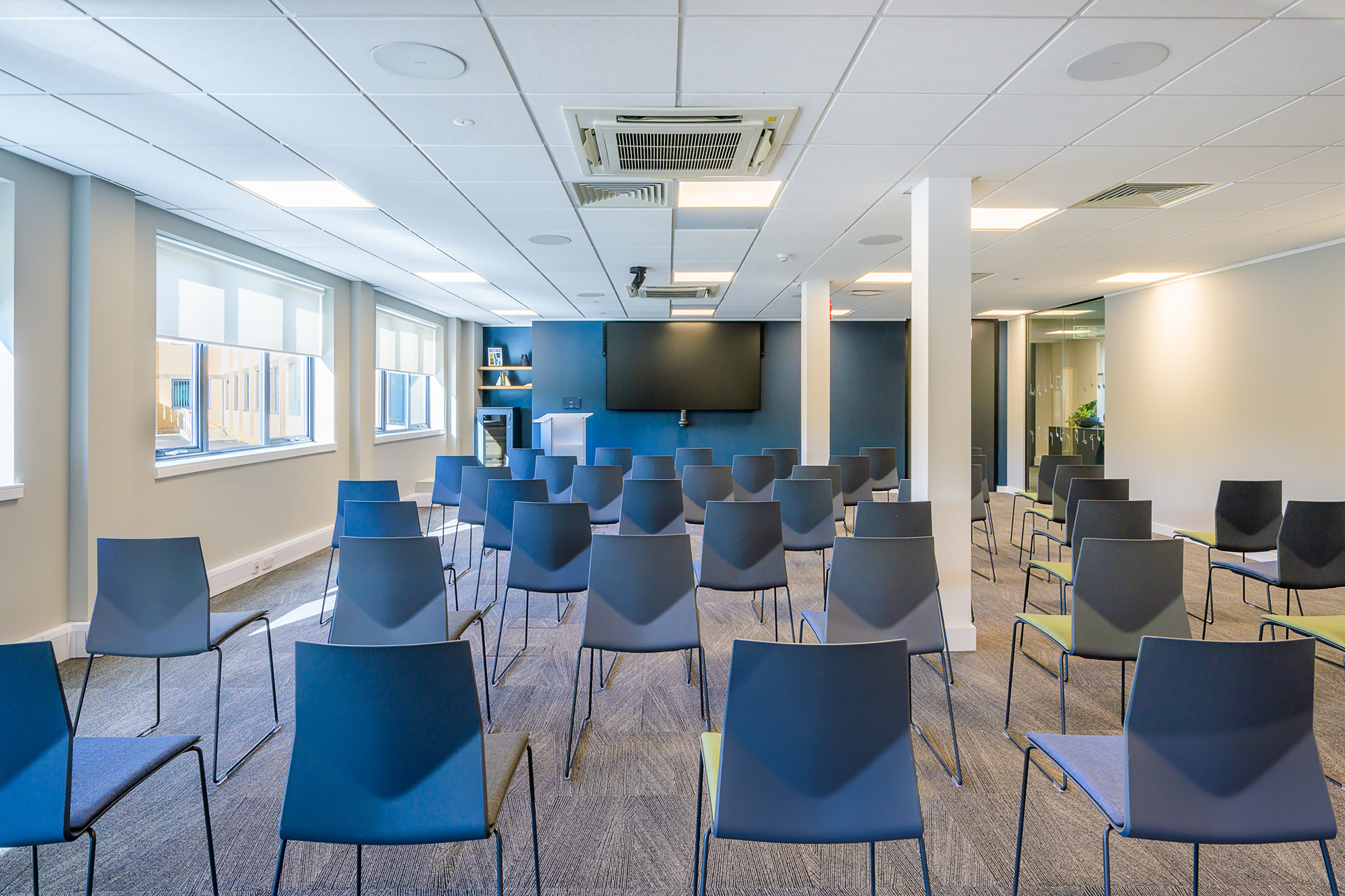Rows of empty blue chairs face a large black screen on a blue wall in a bright conference room with windows and white ceiling panels.