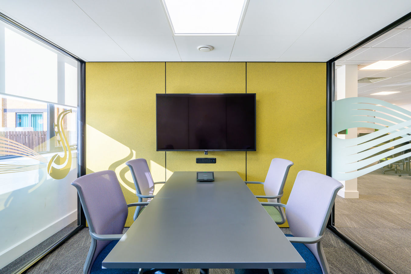 Conference room featuring a central table with chairs surrounding it, a wall-mounted TV screen displaying a blank screen, and large windows with decorative patterns on both sides.