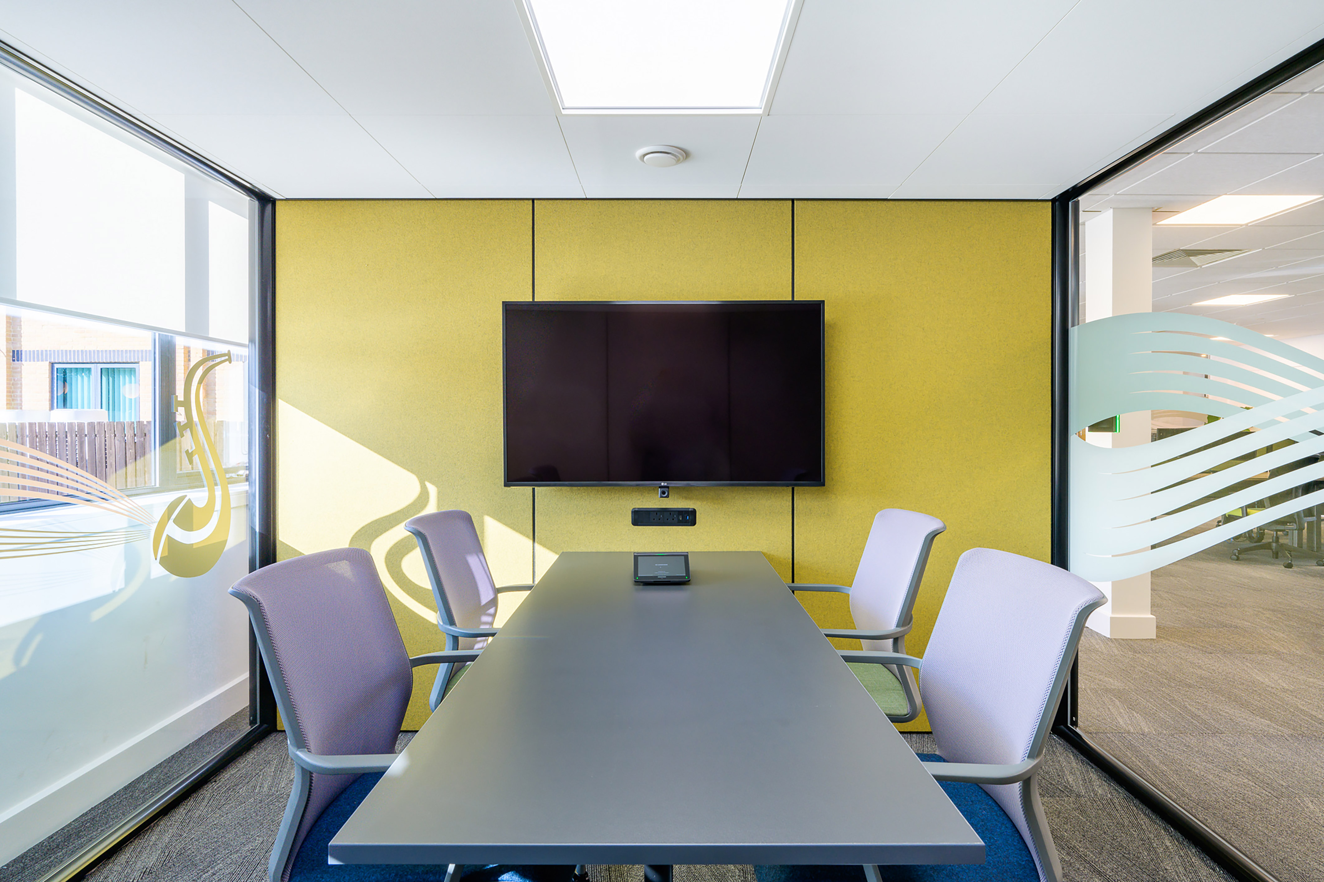Conference room featuring a central table with chairs surrounding it, a wall-mounted TV screen displaying a blank screen, and large windows with decorative patterns on both sides.