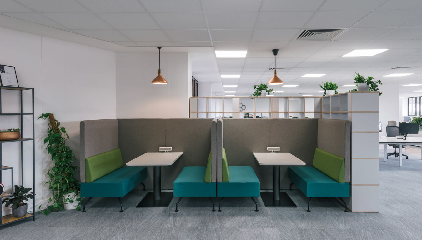 Modern office with cozy booths featuring teal and green seating, accented by pendant lights. Shelves with plants add warmth to the open workspace