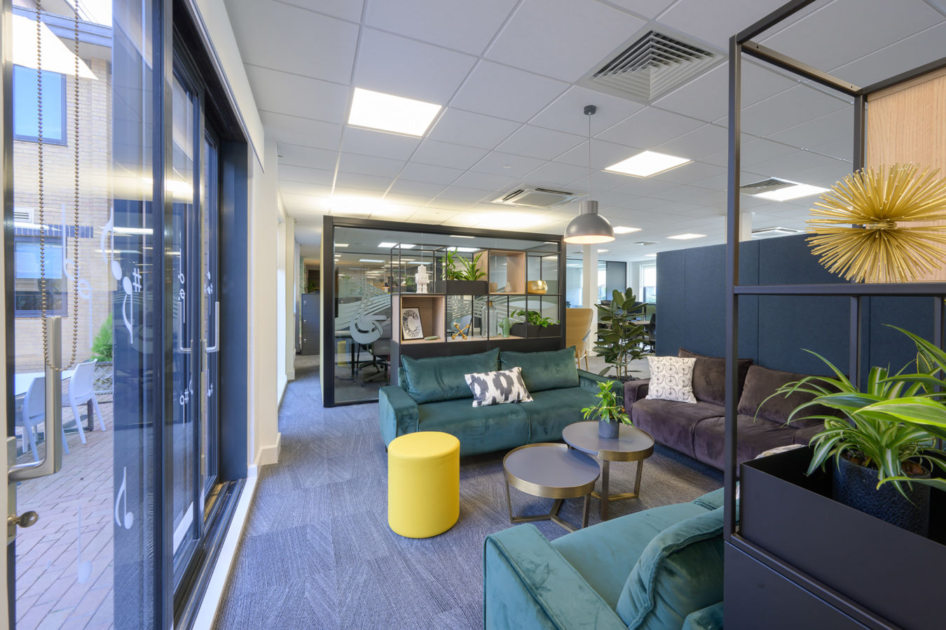 Modern office lounge featuring green and gray sofas with patterned cushions, round tables, and decorative plants; surrounded by glass walls and open workspaces under a ceiling with recessed lighting.