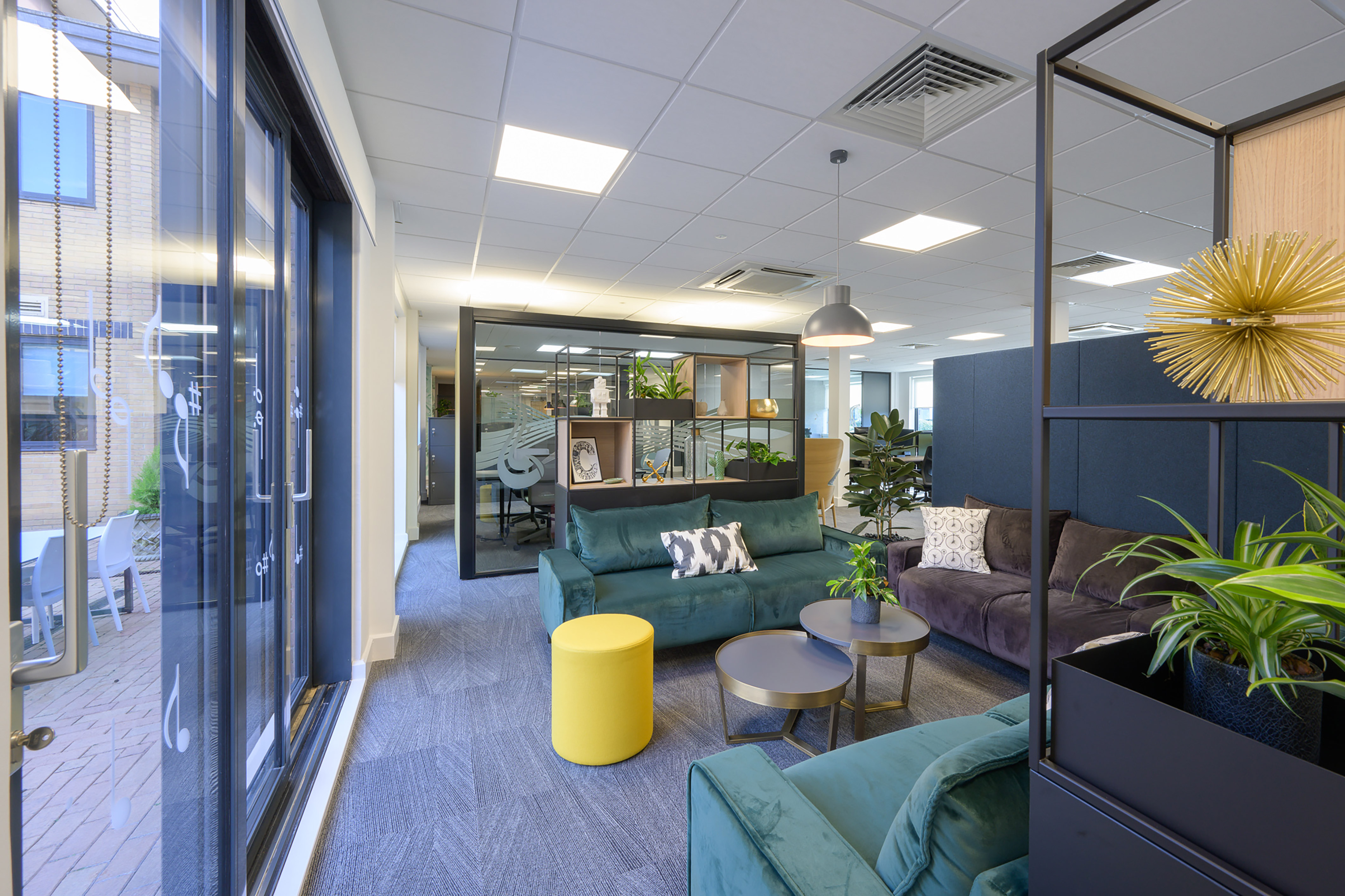 Modern office lounge featuring green and gray sofas with patterned cushions, round tables, and decorative plants; surrounded by glass walls and open workspaces under a ceiling with recessed lighting.