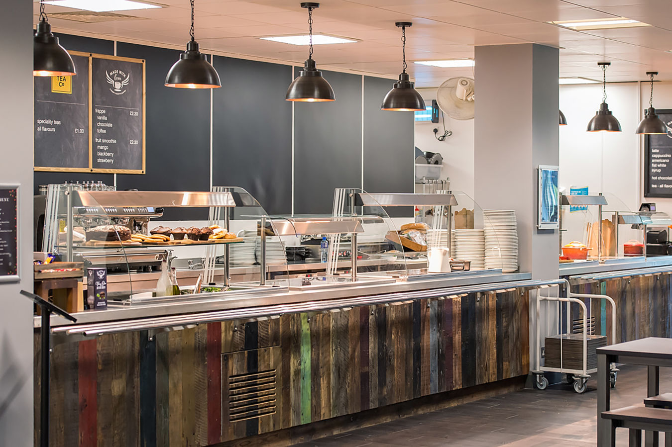 Cafeteria counter displaying various baked goods under glass covers, surrounded by industrial-style decor with pendant lights. Wall menus include “TEA Co” and drink prices like “frappe” at £2.30.