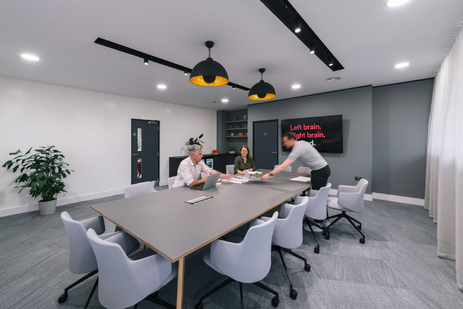 Modern conference room with a large table and eight chairs. Three people are engaged in discussion. A screen displays "Left brain. Right brain." Bright lighting and plant decor create an inviting atmosphere