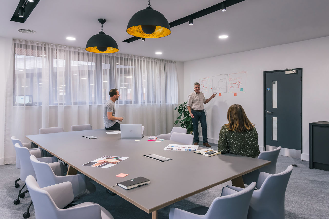 Modern meeting room with pendant lighting, a large conference table, and people engaged in a discussion near a whiteboard with notes.