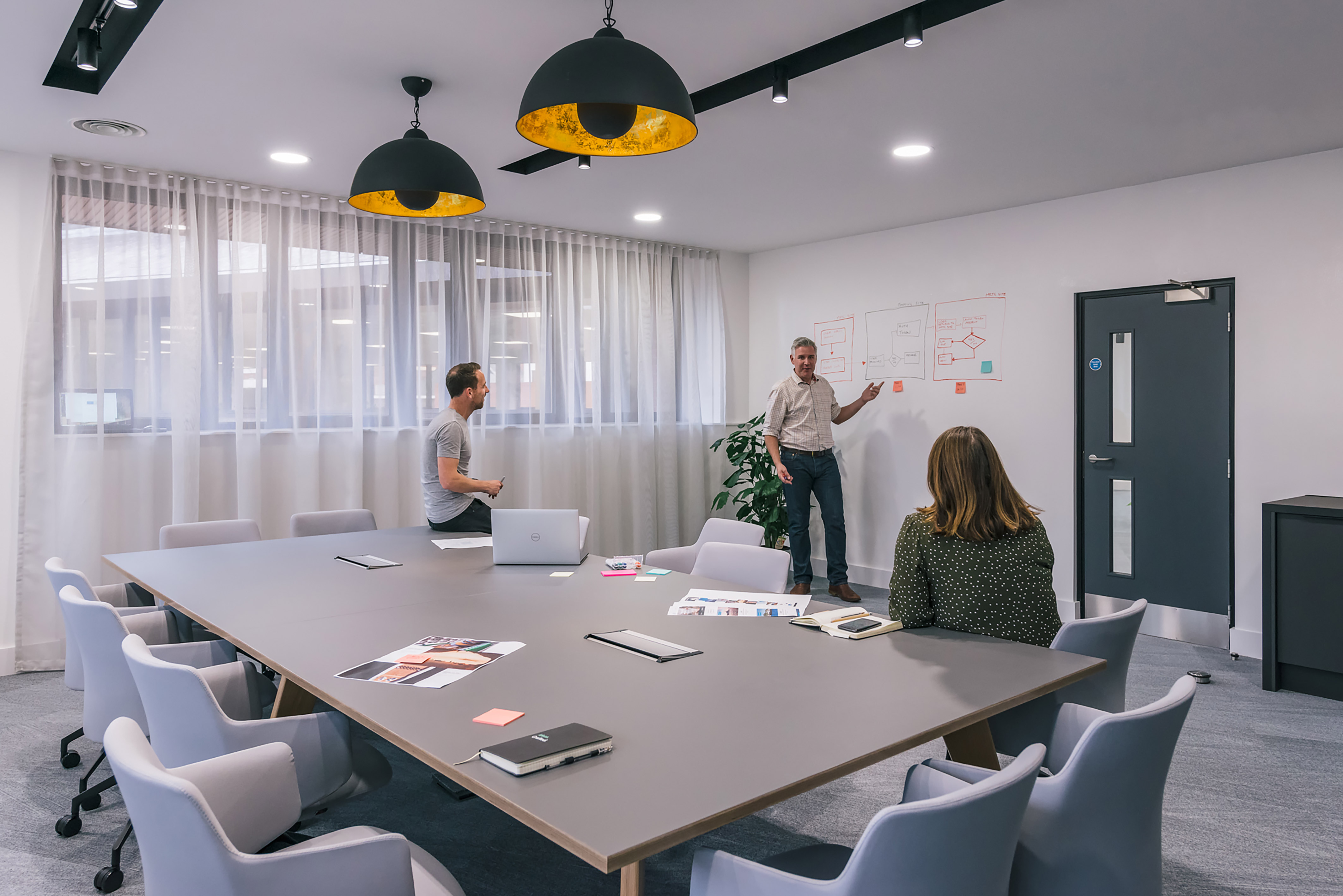 Modern meeting room with pendant lighting, a large conference table, and people engaged in a discussion near a whiteboard with notes.