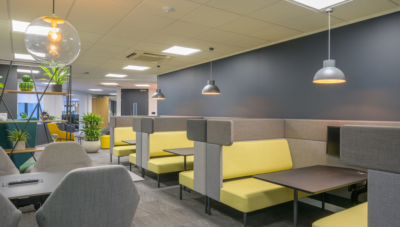 Modern office lounge with gray and yellow booth seating, gray walls, and hanging lights. Shelves with plants add a fresh touch. Bright, inviting atmosphere