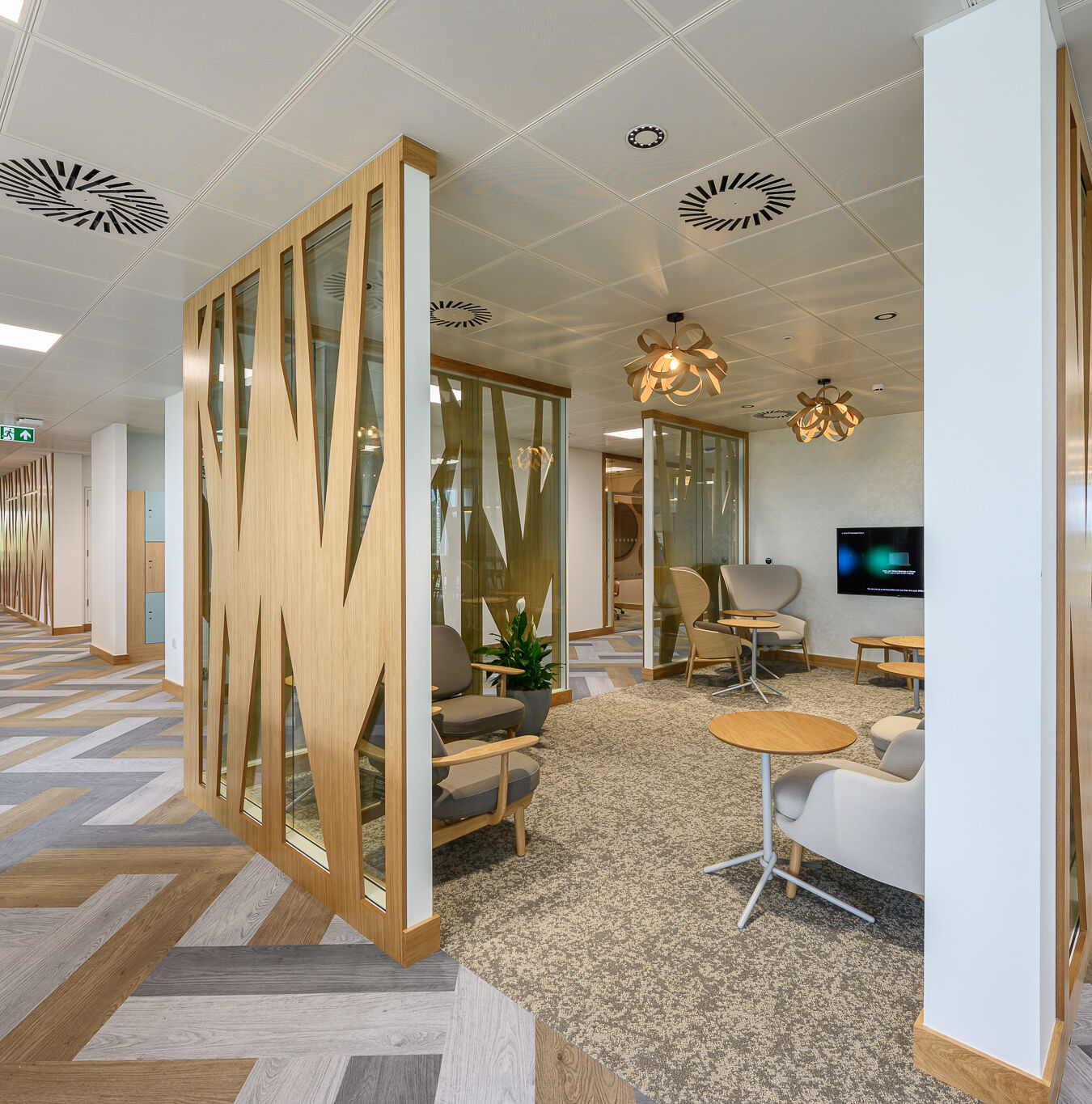 Modern office interior with wood accents and glass walls, featuring cozy seating, contemporary pendant lights, patterned flooring, and a TV. Bright and inviting