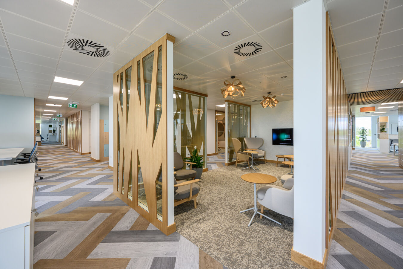 Modern office interior with wood accents and glass walls, featuring cozy seating, contemporary pendant lights, patterned flooring, and a TV. Bright and inviting