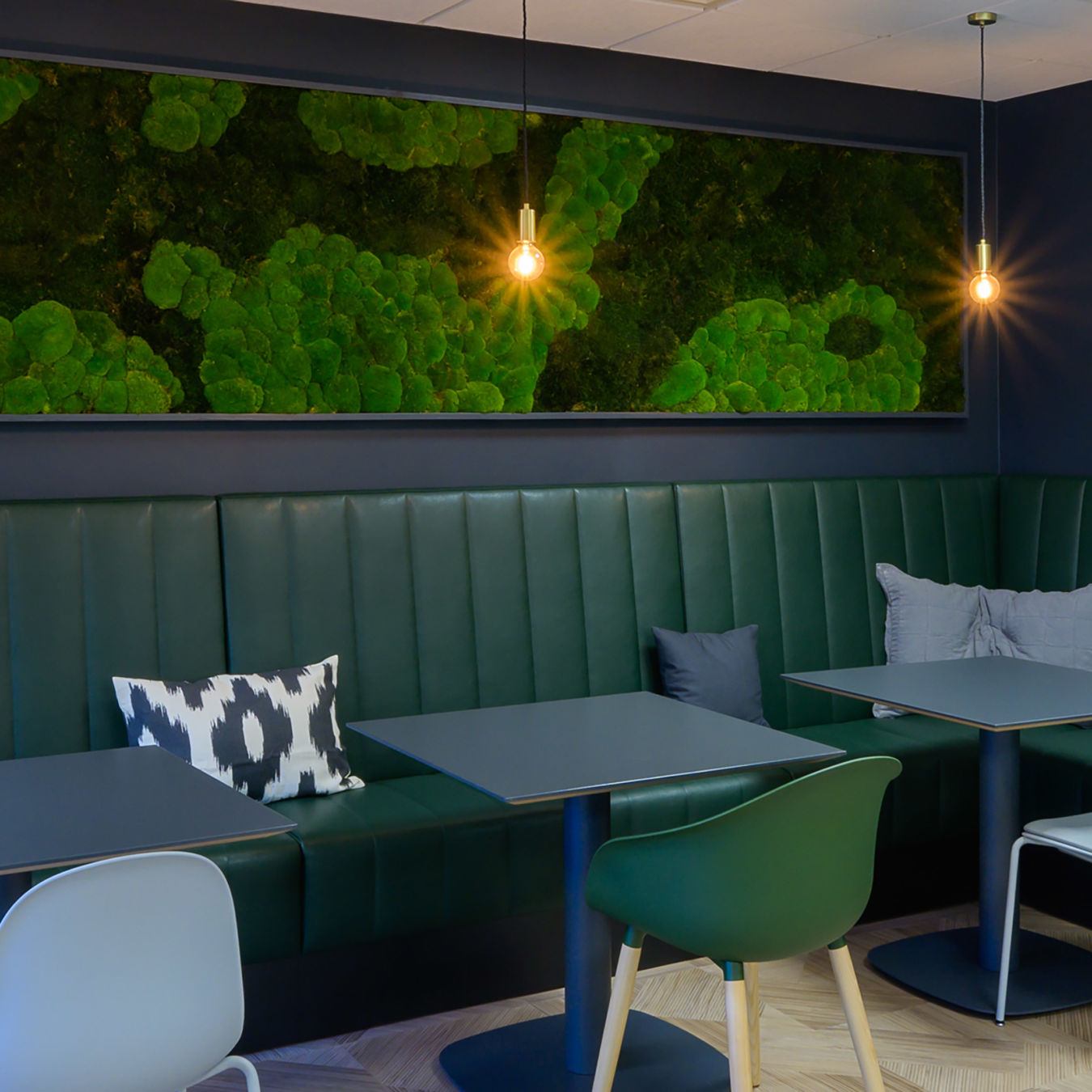 dark green leather booth seating and two sleek tables. Warm pendant lights hang above, casting a cozy glow. A vibrant green textured artwork adorns the deep blue wall, enhancing the calming atmosphere