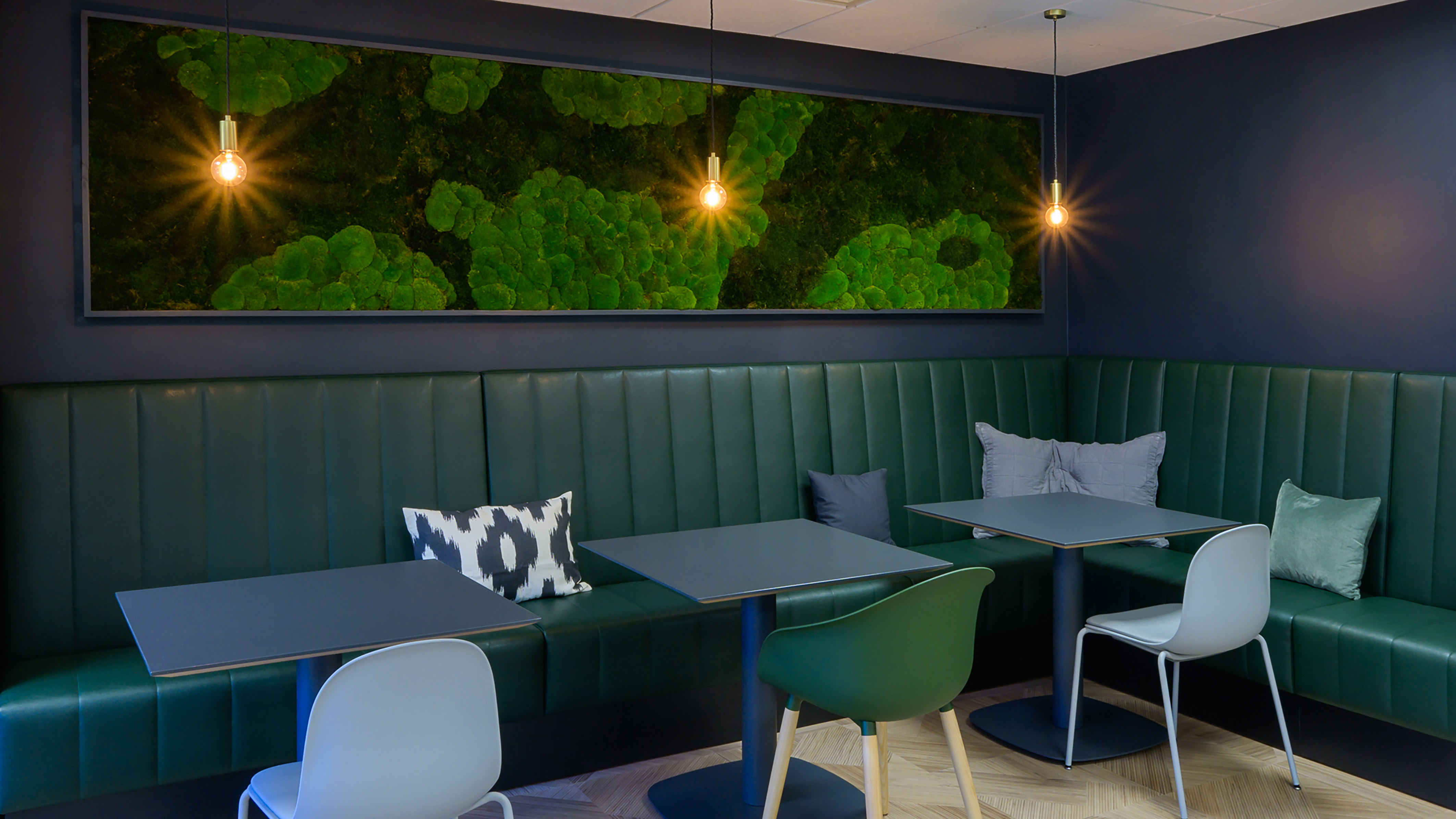 dark green leather booth seating and two sleek tables. Warm pendant lights hang above, casting a cozy glow. A vibrant green textured artwork adorns the deep blue wall, enhancing the calming atmosphere