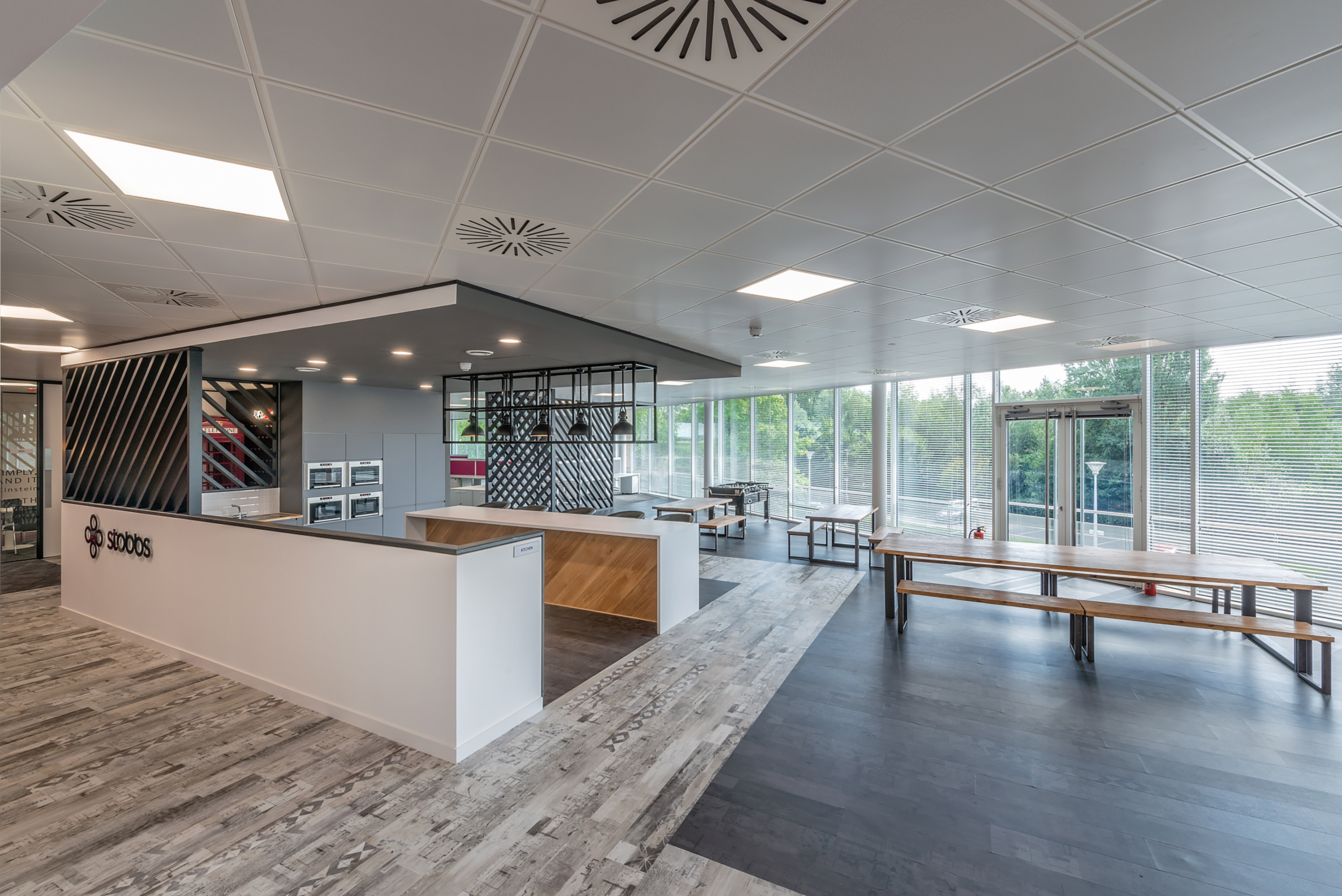 Modern, spacious office break room with a sleek kitchen area, long wooden tables, large windows with blinds, and gray flooring. Bright and airy ambiance