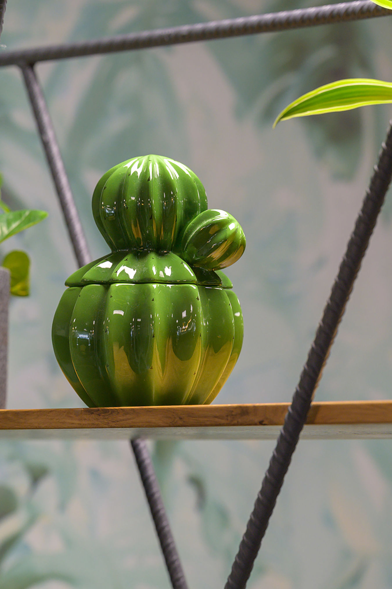 Green ceramic cactus-shaped container sits on a wooden shelf amid a blurred, abstract leafy background.