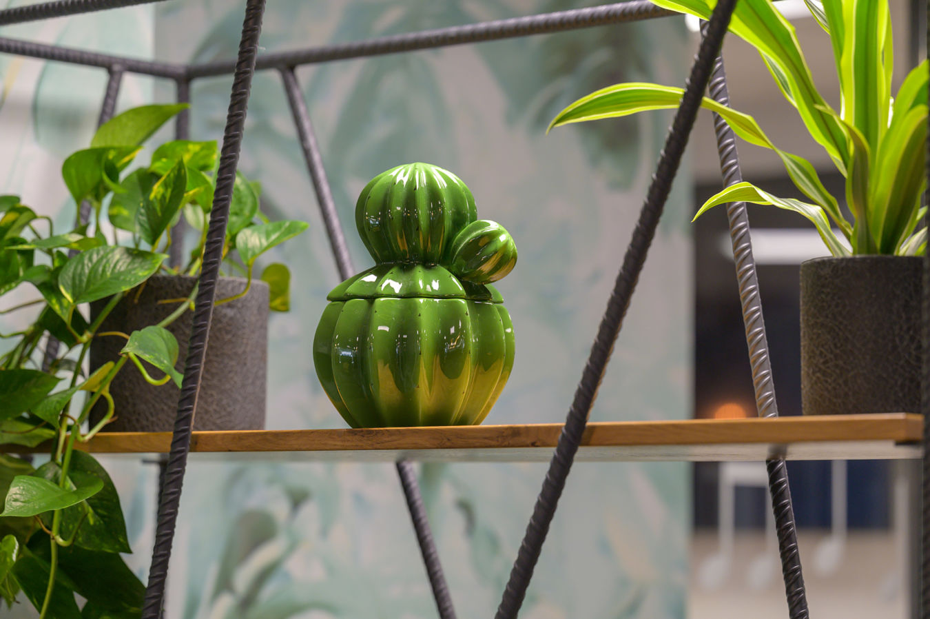 Green ceramic cactus-shaped container sits on a wooden shelf amid a blurred, abstract leafy background.