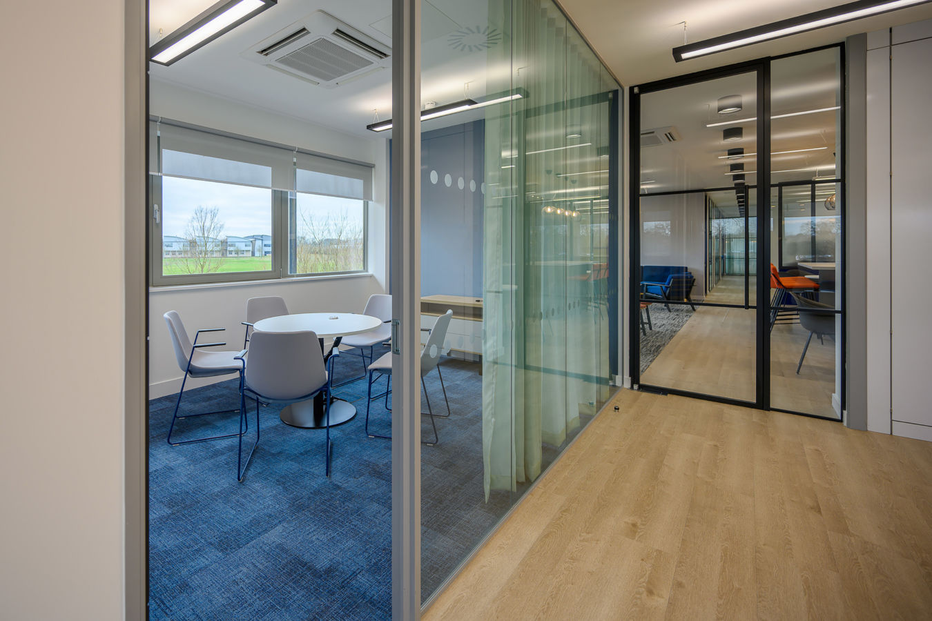 Modern meeting room with round table and four chairs, separated by glass walls. The space features blue carpet, light wood flooring, and large windows overlooking greenery. Ceiling lighting is visible.