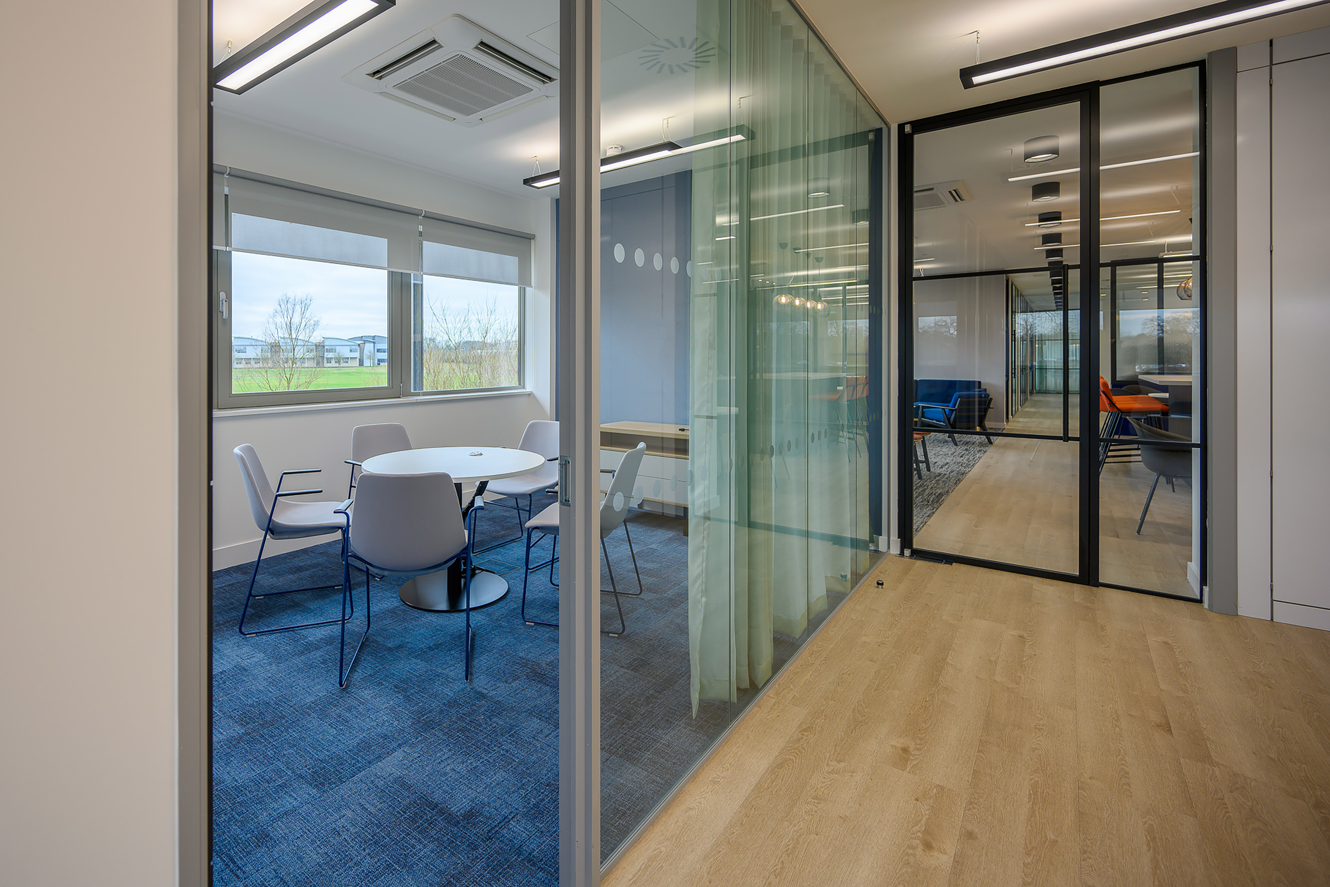 Modern meeting room with round table and four chairs, separated by glass walls. The space features blue carpet, light wood flooring, and large windows overlooking greenery. Ceiling lighting is visible.
