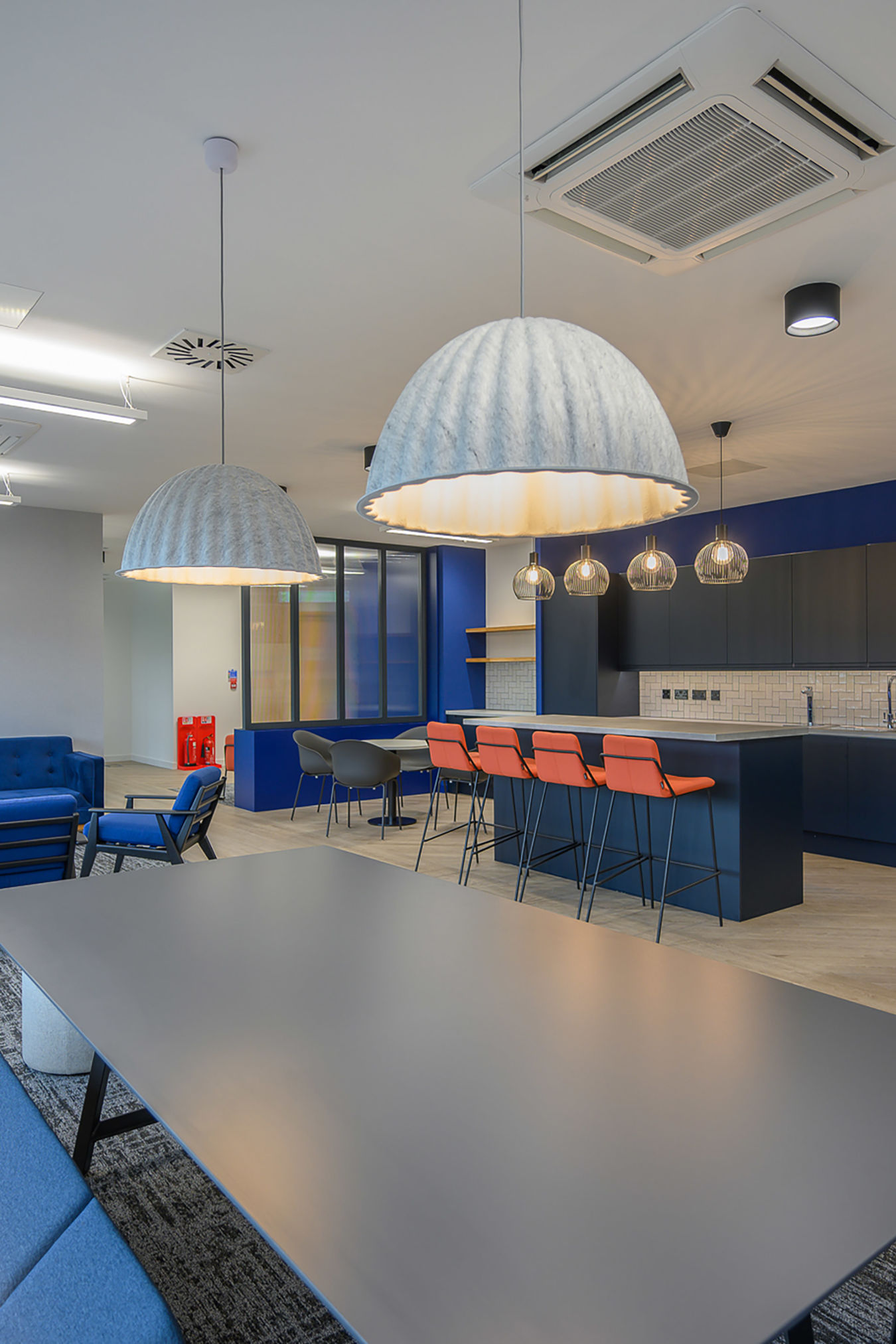 Large pendant lights hang over a sleek, modern office break room featuring a black counter with orange stools and blue furniture, illuminated by soft ambient lighting.