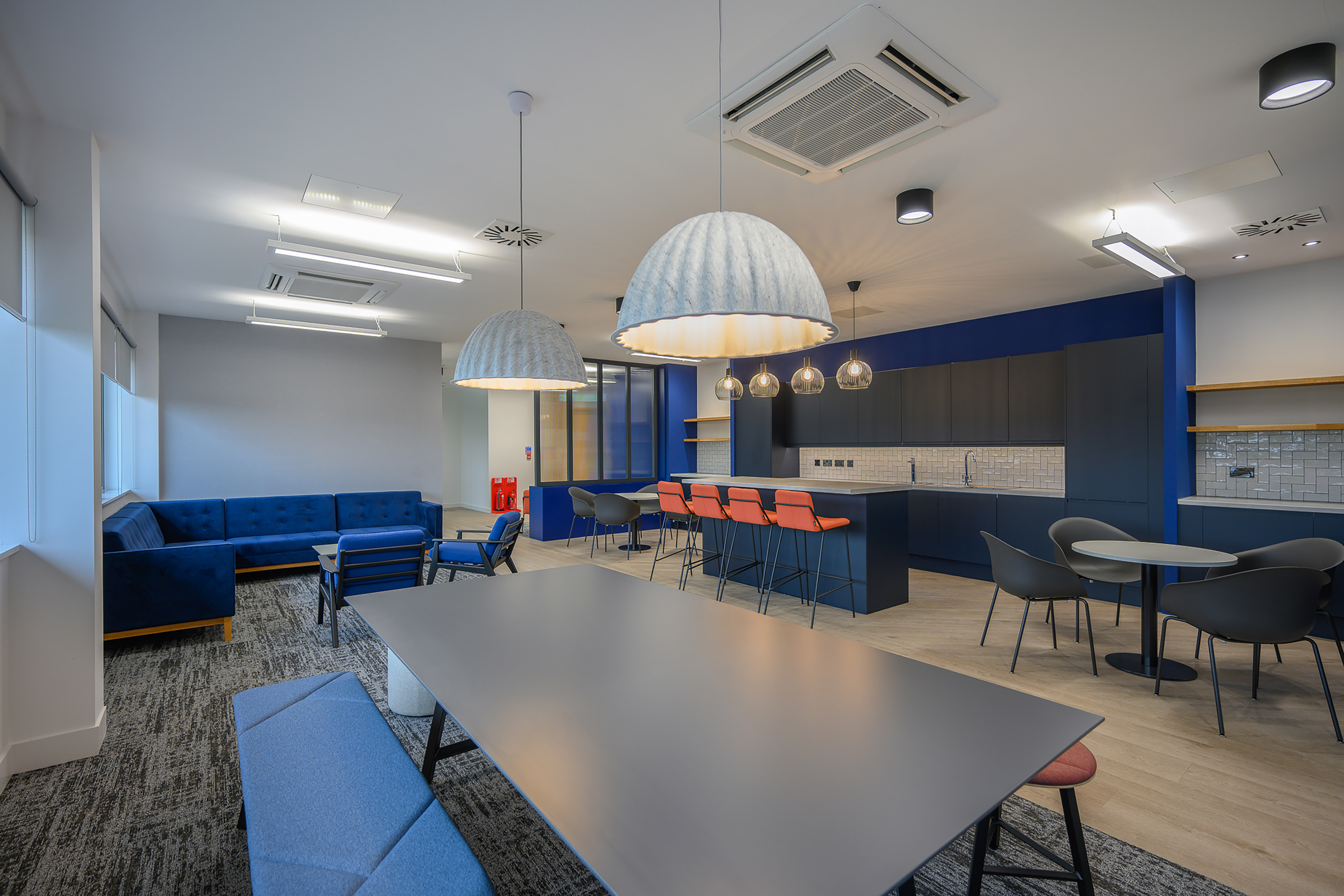 Large pendant lights hang over a sleek, modern office break room featuring a black counter with orange stools and blue furniture, illuminated by soft ambient lighting.