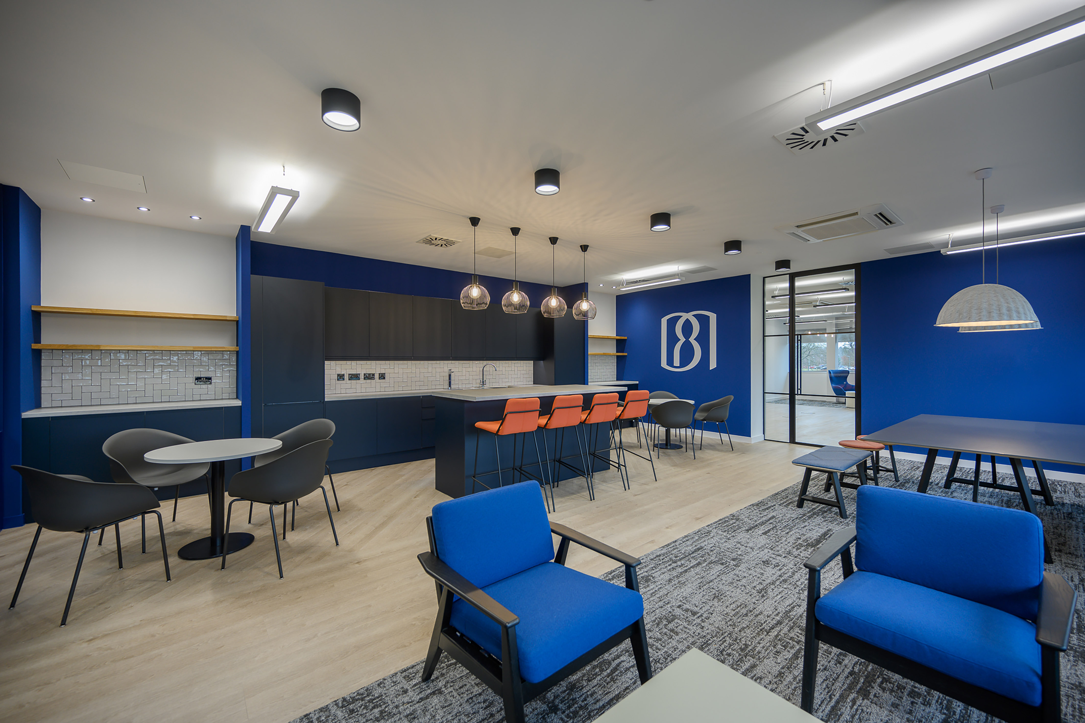 Modern office lounge features blue armchairs, a round table, and a kitchenette with orange stools. Pendant lights hang above a counter. A blue wall displays an abstract "8" logo.