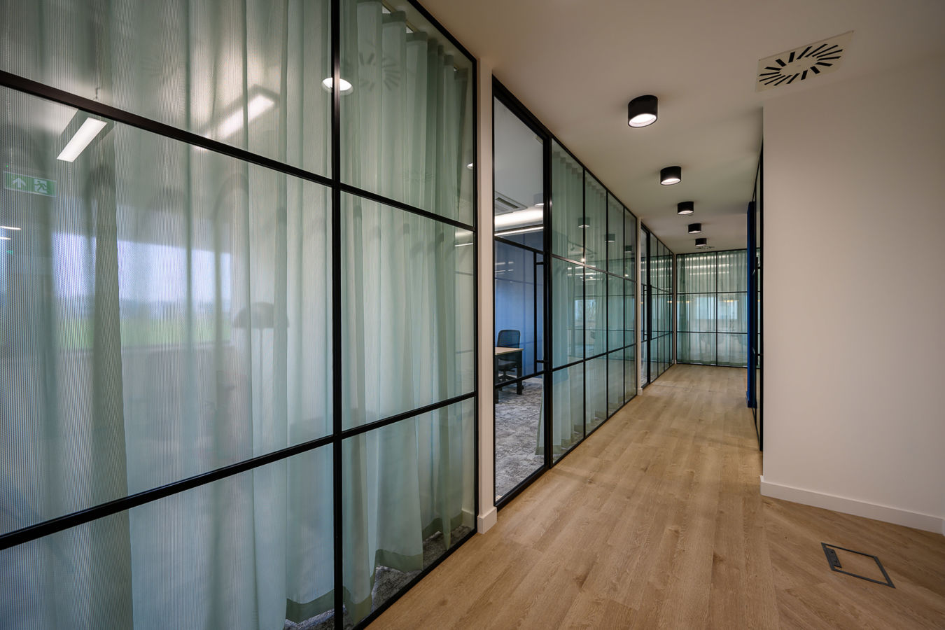 Glass-walled corridor with sheer curtains leads to an office room; ceiling lights illuminate the wood flooring. An exit sign is visible at the end of the hallway.