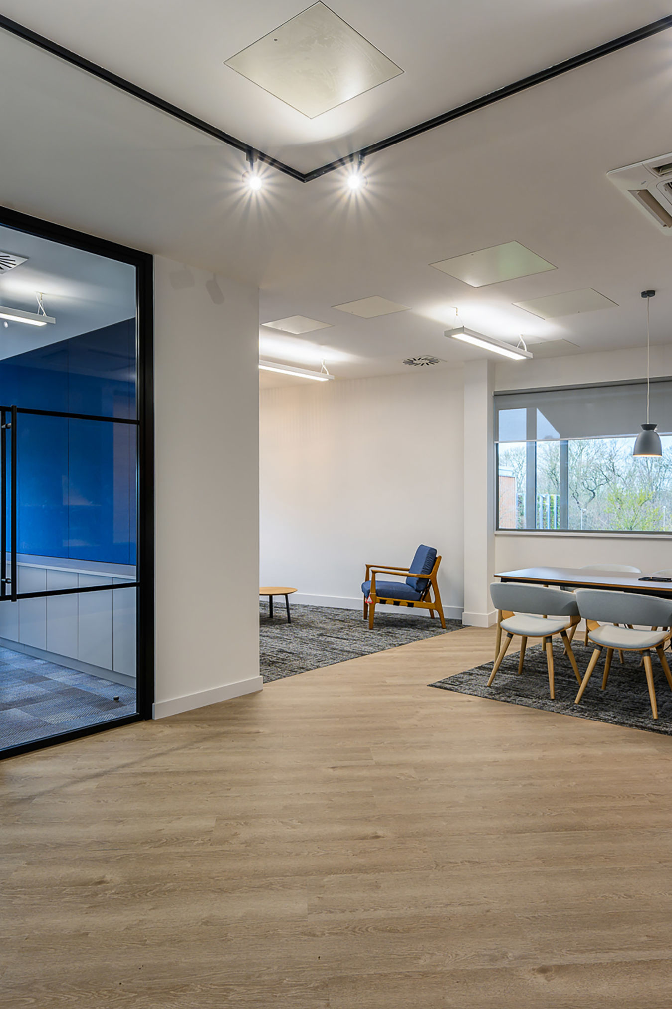 Modern office interior with wooden flooring includes a blue chair, a round table, and five white chairs around a rectangular table under overhead lights, near a large window.