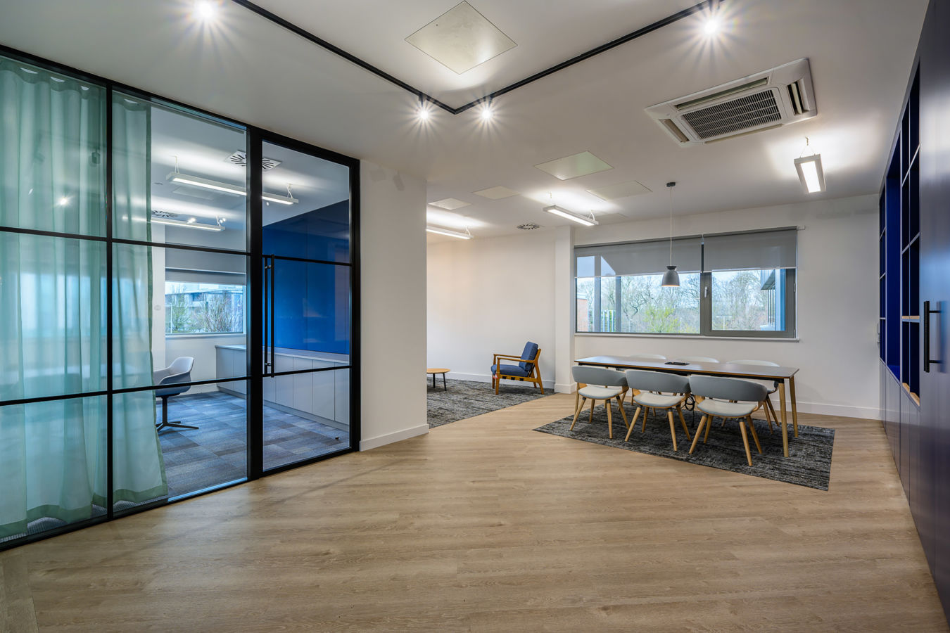 Modern office interior with wooden flooring includes a blue chair, a round table, and five white chairs around a rectangular table under overhead lights, near a large window.
