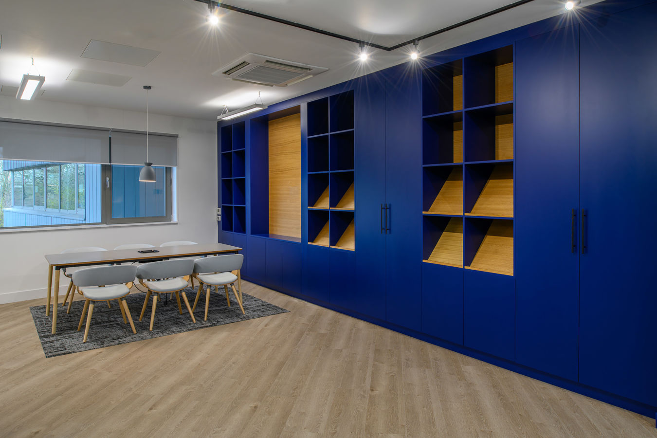 Office meeting room with a wooden table and six white chairs, adjacent to large blue shelves. The room features wooden flooring and a window with a view of greenery.