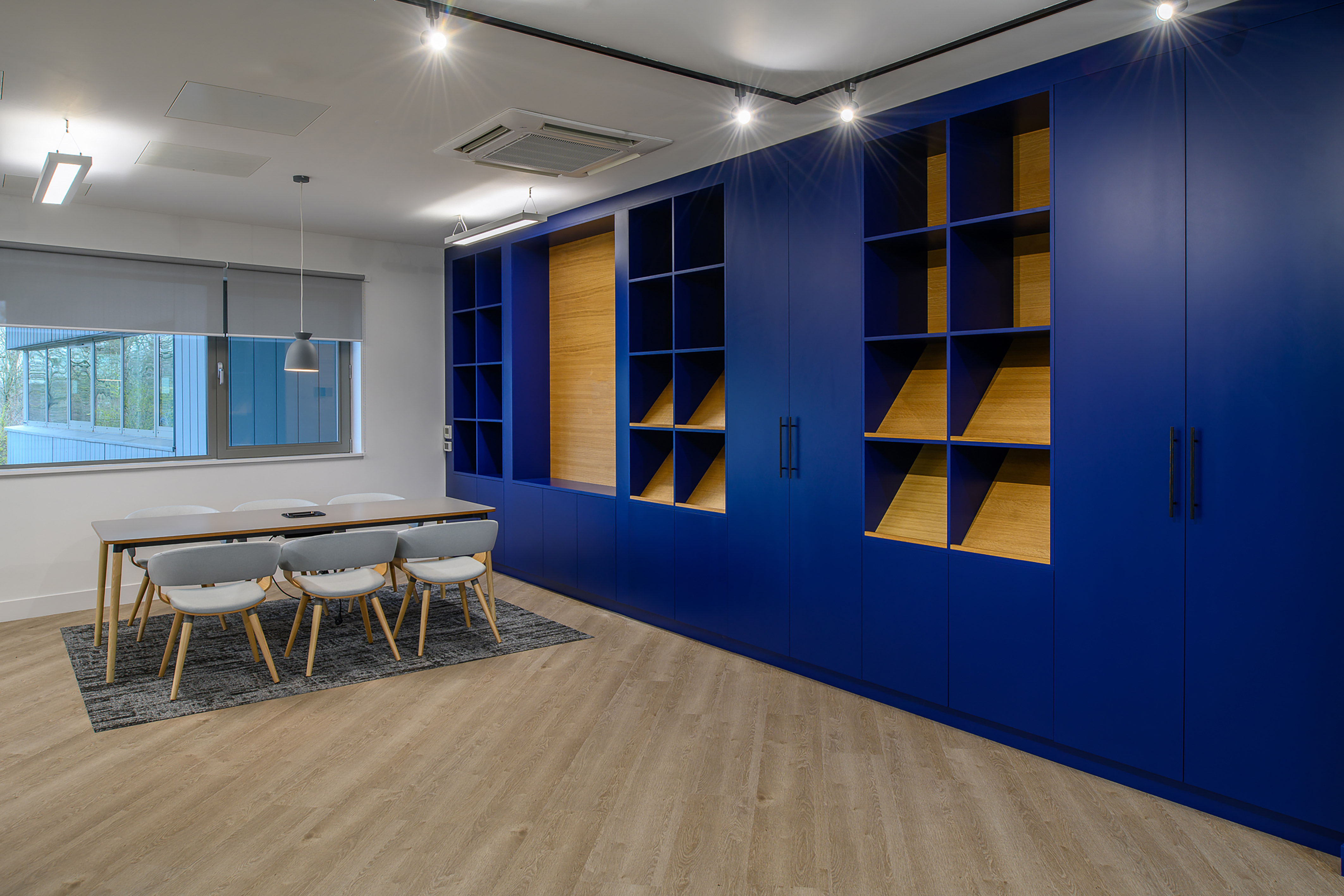 Office meeting room with a wooden table and six white chairs, adjacent to large blue shelves. The room features wooden flooring and a window with a view of greenery.