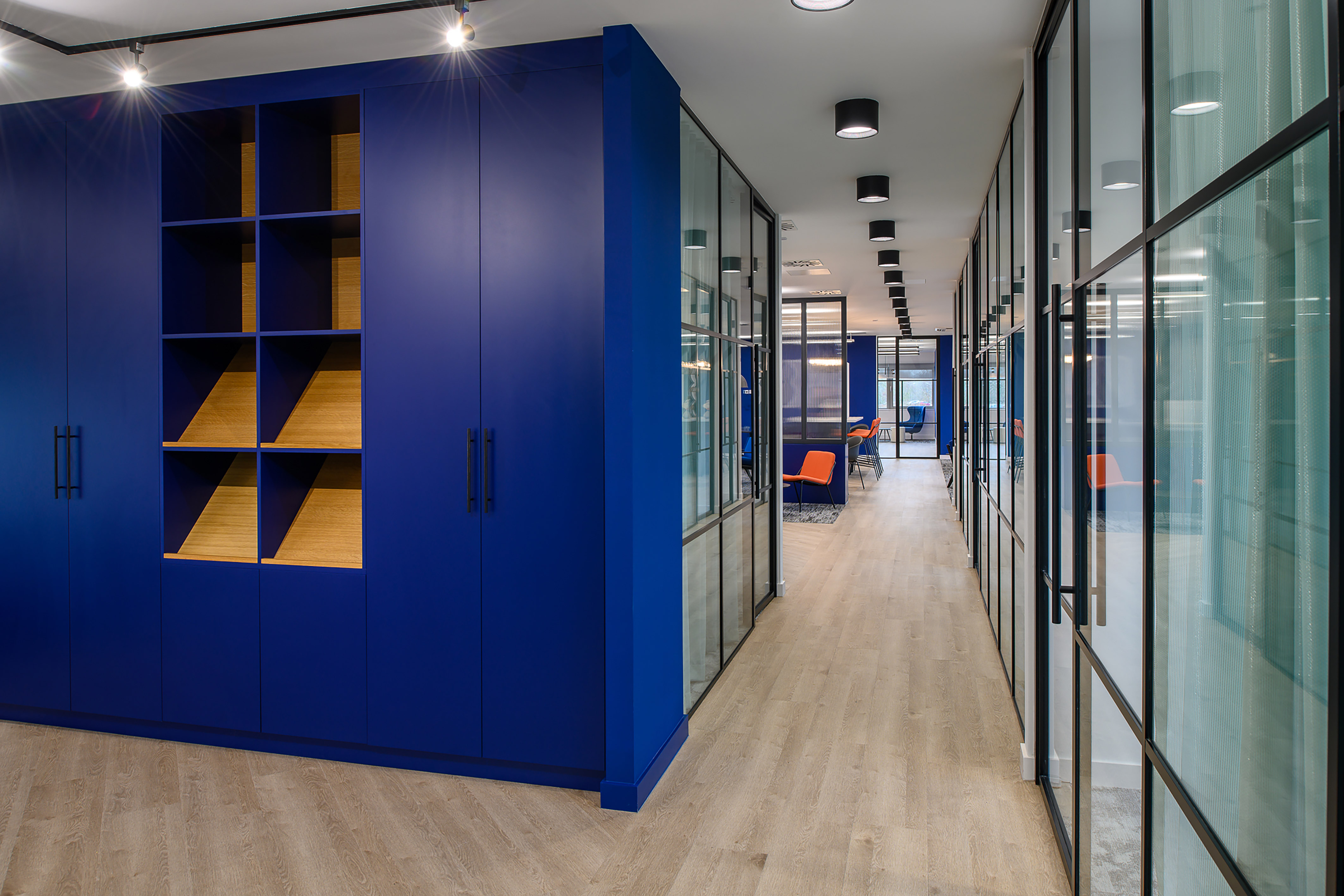 Blue cabinet features empty wooden shelves in an office hallway with glass walls and orange chairs, illuminated by overhead lights on a wooden floor.