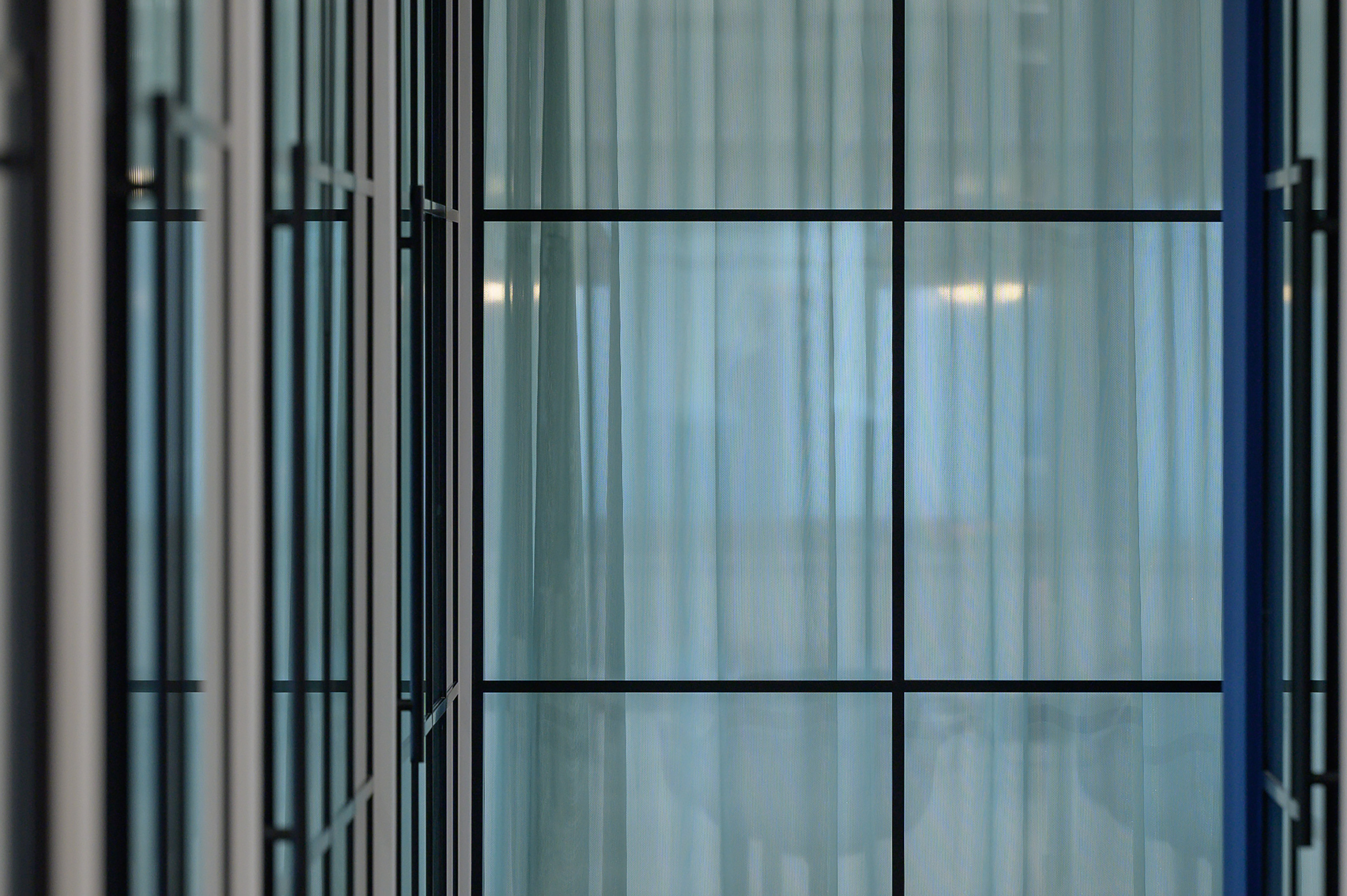 Glass panels form a reflective corridor with parallel black frames, creating a modern and minimalist architectural design with diffused light filtering through delicate curtains.