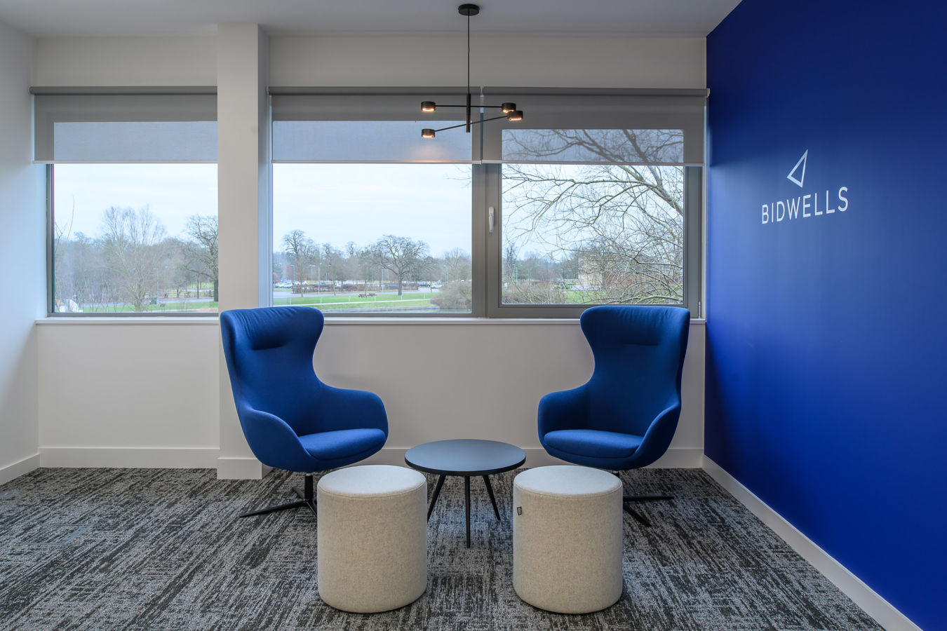 Two blue armchairs face each other with a round black table in between, set on a patterned carpet in a room with a large window overlooking a park.