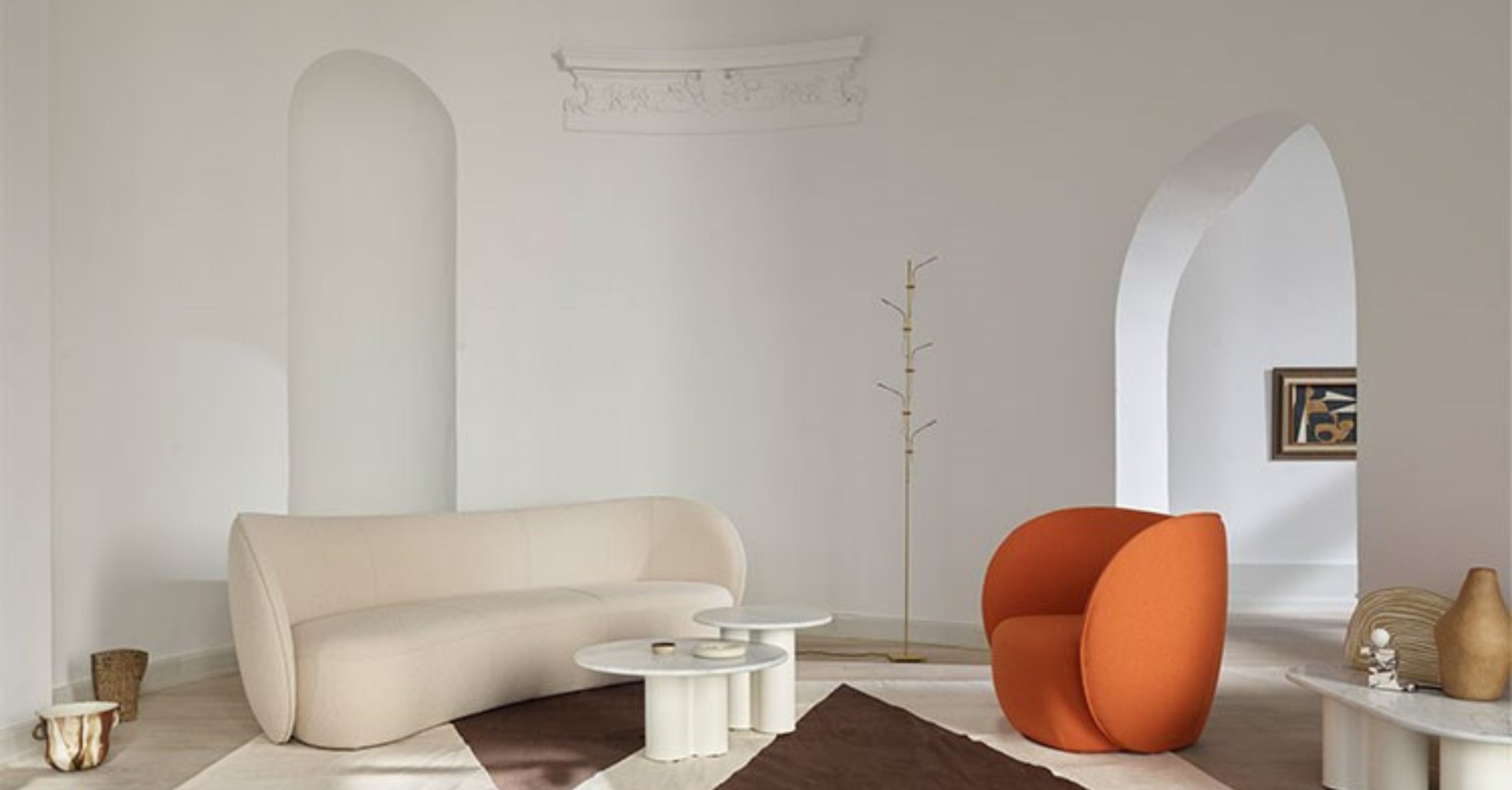 Minimalist lounge with a curved beige sofa, round nesting tables, bold orange armchair, and sculptural gold coat stand