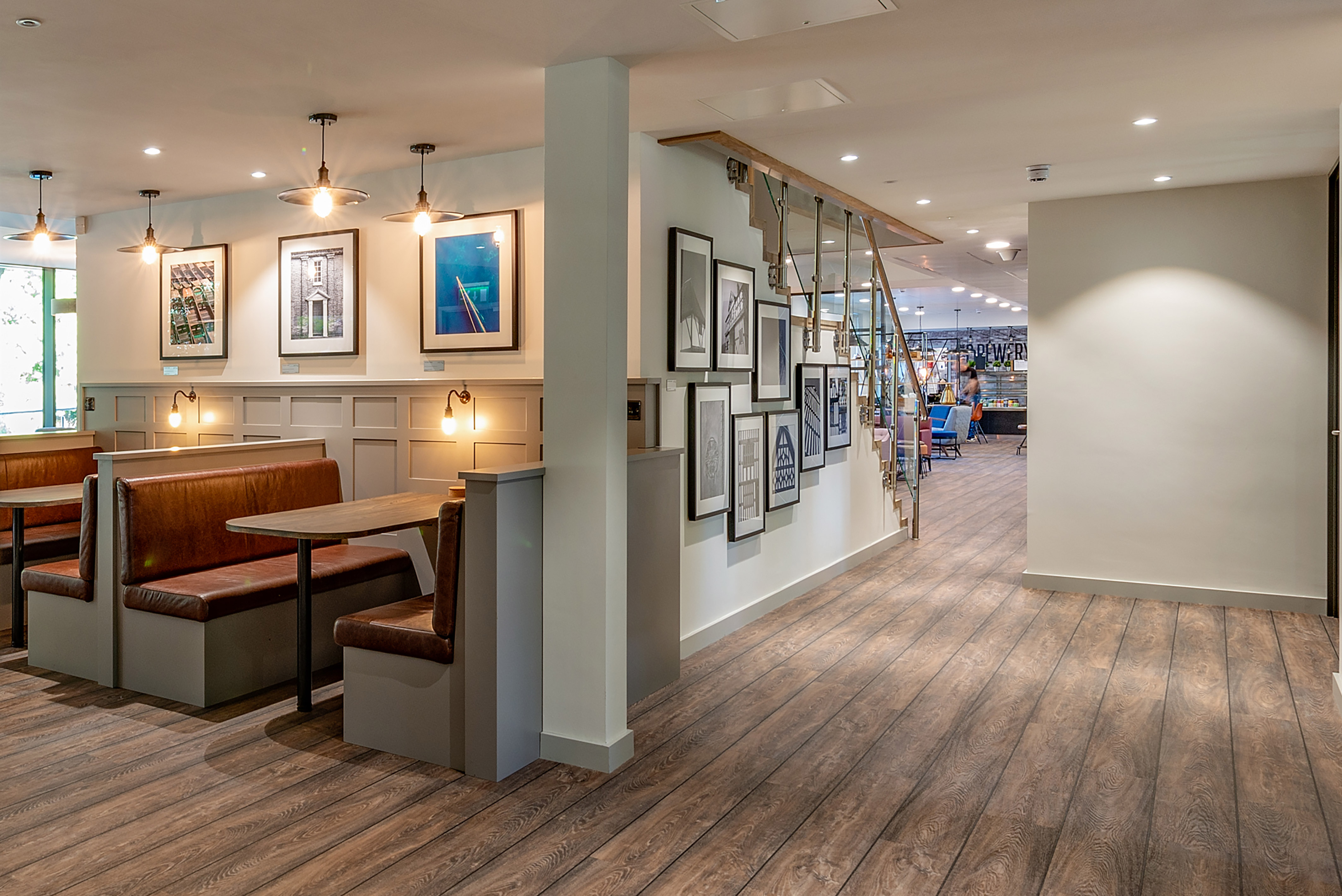 Warm, modern cafe interior with brown leather booth seating on the left, framed art on walls, wooden floors, and a view into a busy dining area. Cozy ambiance