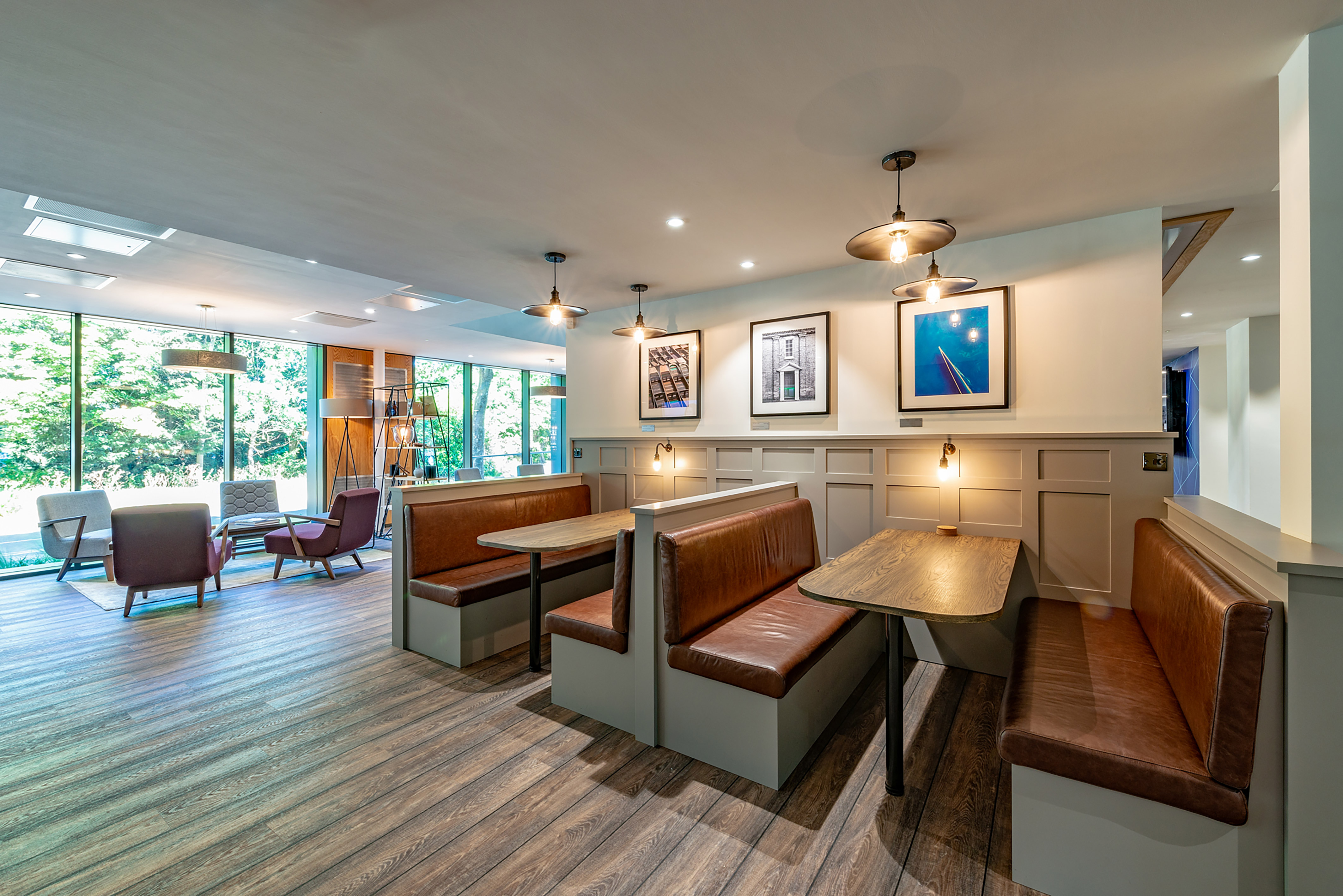 Booths with brown leather seating and wooden tables sit under framed art and pendant lights in a bright cafe area. Large windows reveal a green outdoor view.