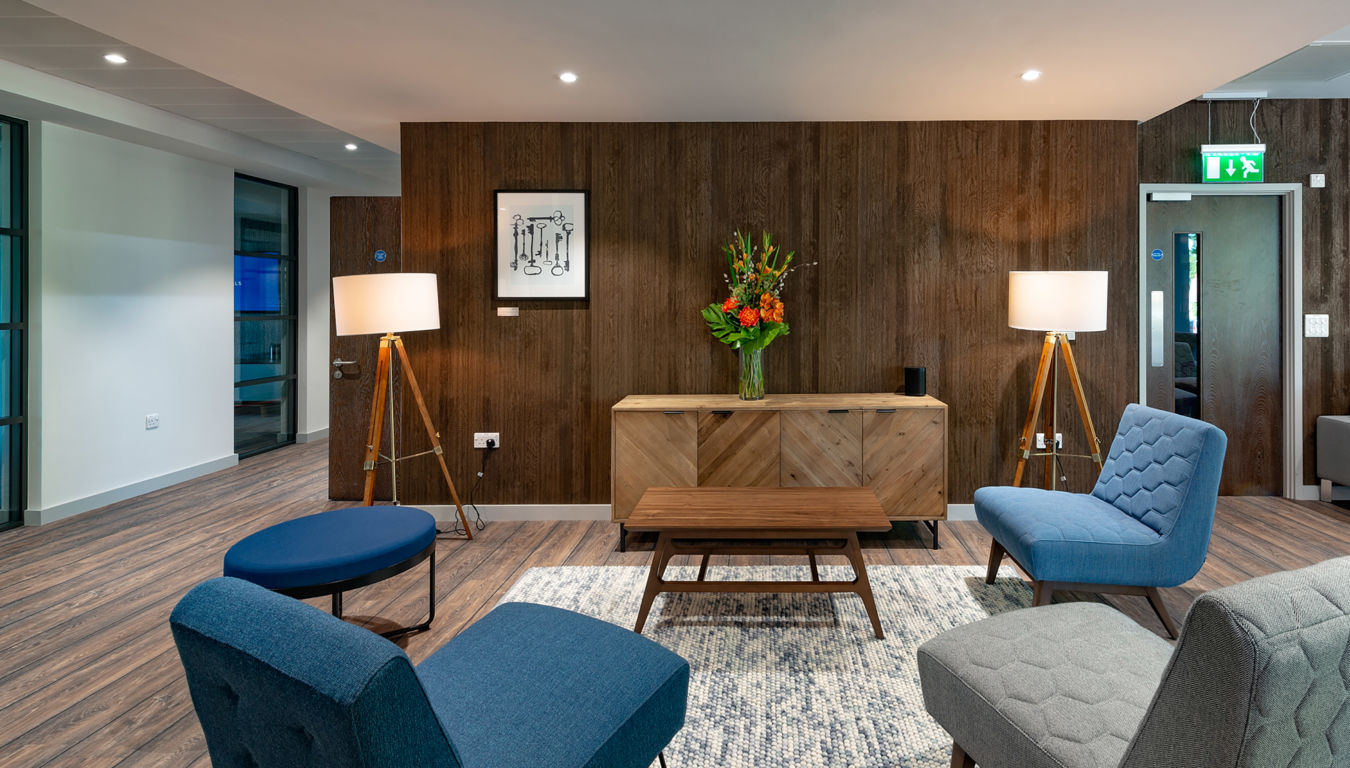 Modern lounge with mid-century style chairs in blue and gray, wooden panel wall, floor lamps, vibrant flower arrangement, and soft lighting. Cozy ambiance