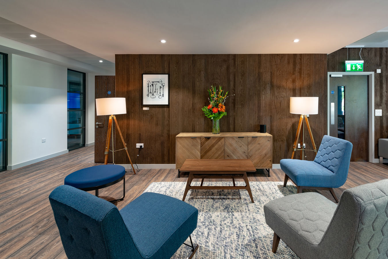 Modern lounge with mid-century style chairs in blue and gray, wooden panel wall, floor lamps, vibrant flower arrangement, and soft lighting. Cozy ambiance