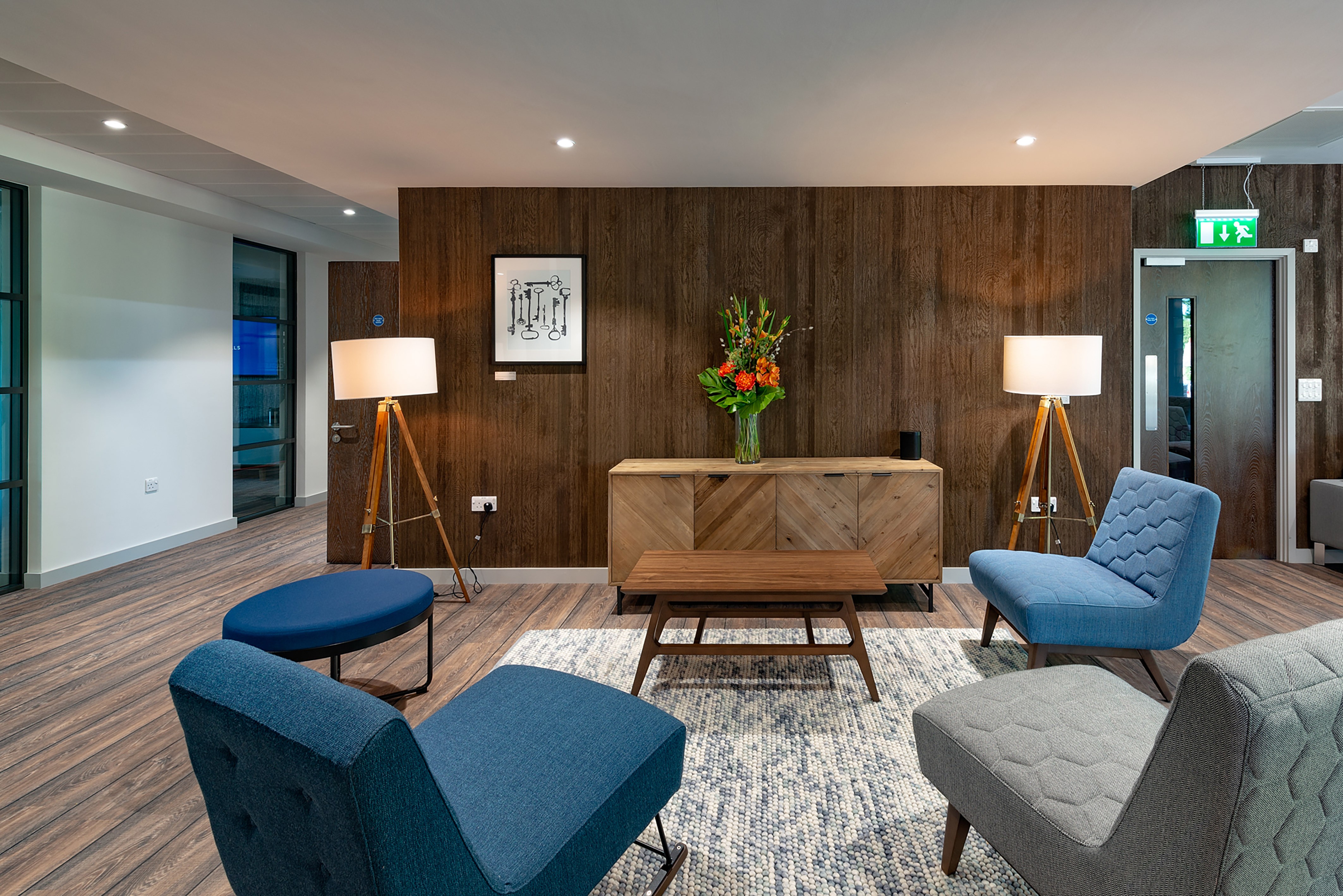 Modern lounge with mid-century style chairs in blue and gray, wooden panel wall, floor lamps, vibrant flower arrangement, and soft lighting. Cozy ambiance