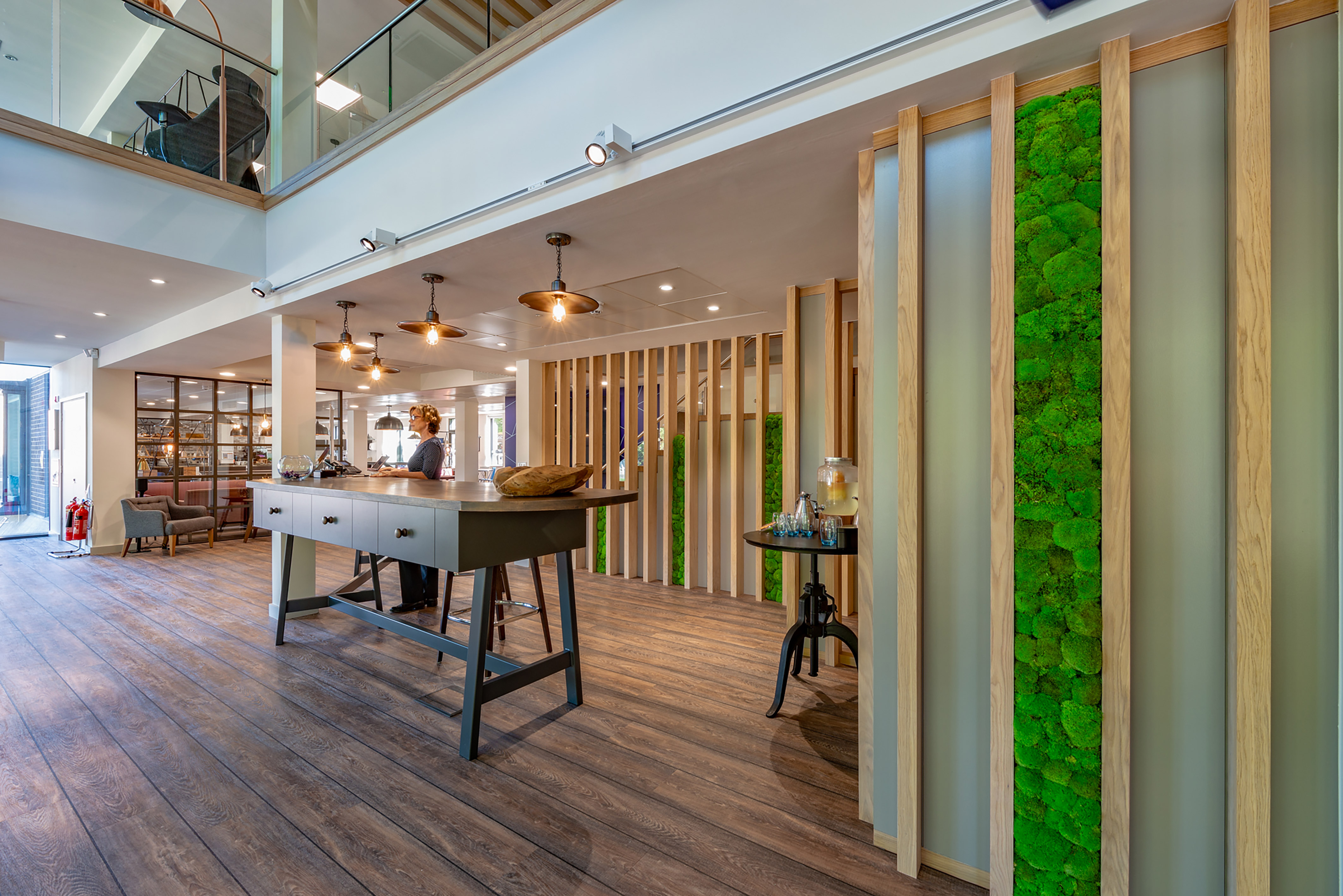 A modern reception area features a person standing at a central table under hanging lights, surrounded by wooden partitions and green moss wall decor.